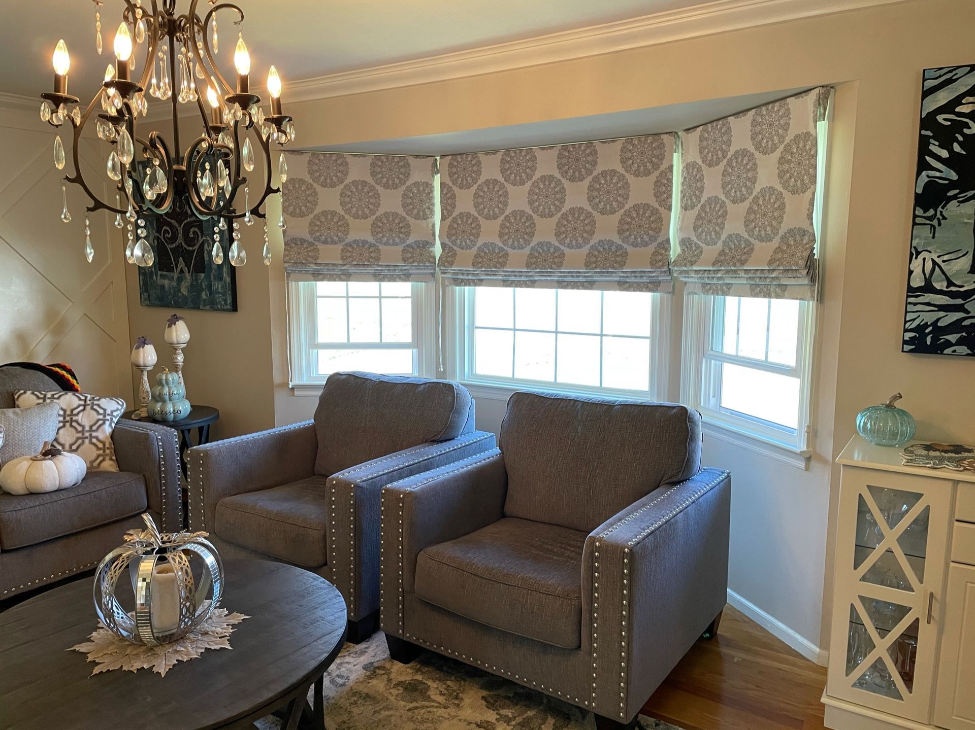 Living room with two gray armchairs facing a bay window with patterned shades; chandelier.