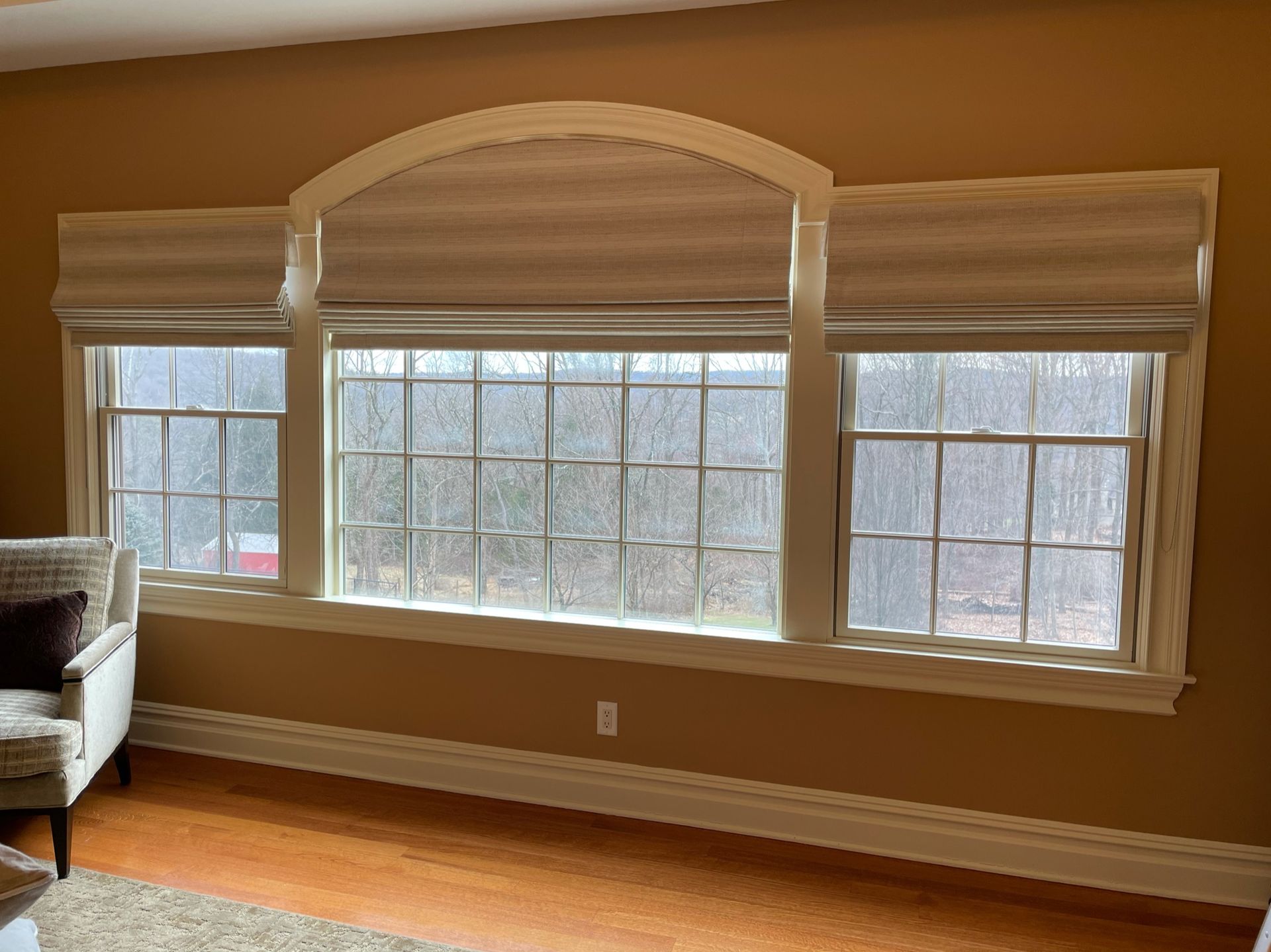Three windows with Roman shades and arch detail on a tan wall, with a chair visible on the left.