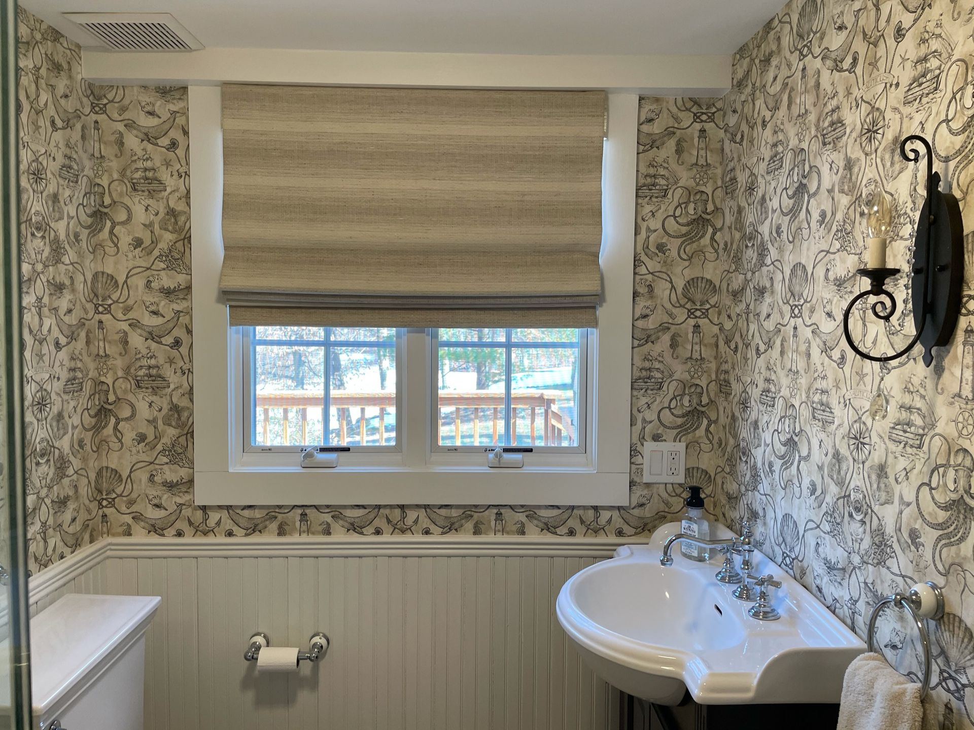 Bathroom with patterned wallpaper, window with tan shade, white sink, and black sconce.