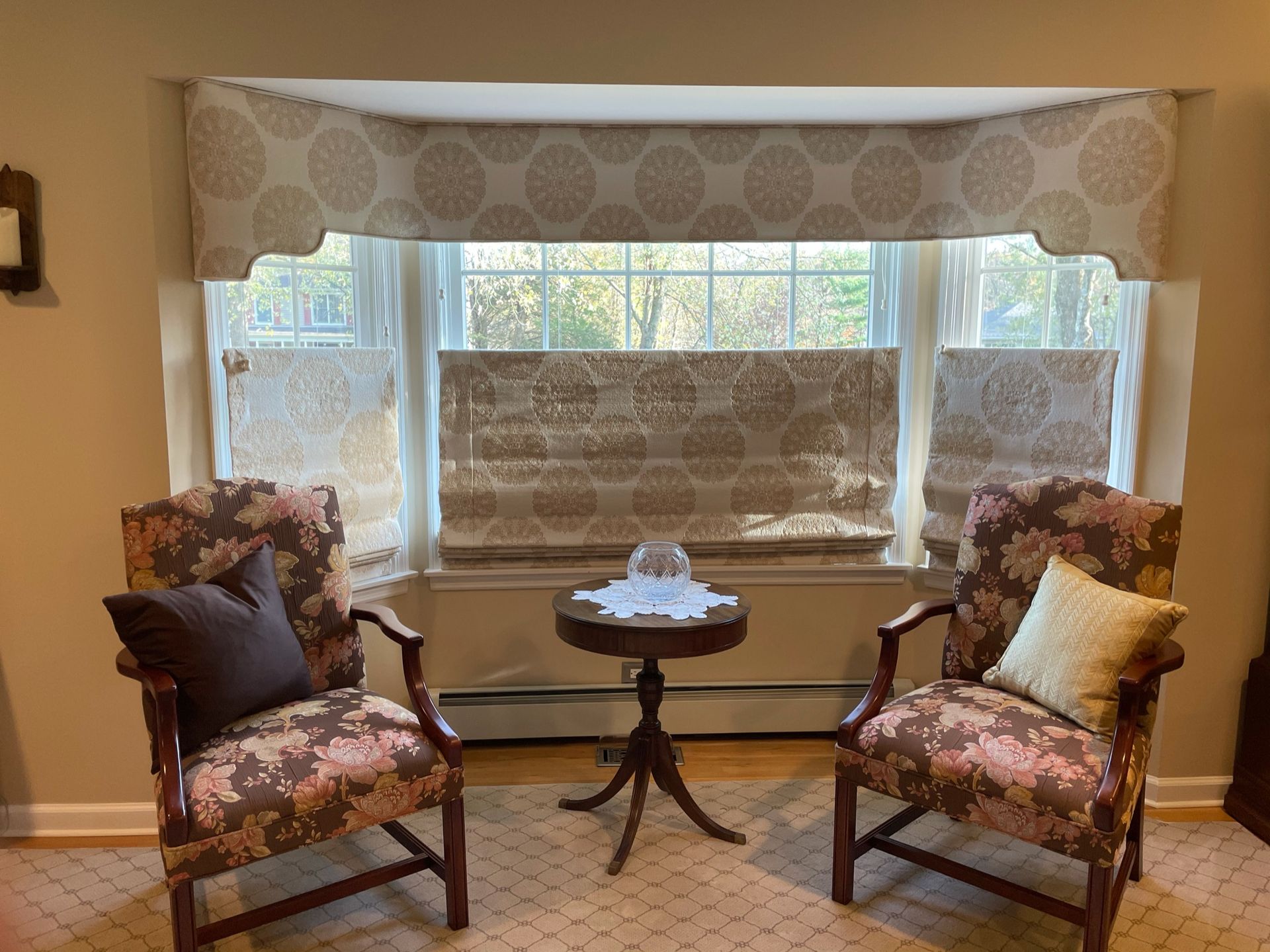 Two floral armchairs face a small table in front of a bay window with patterned shades and valance.