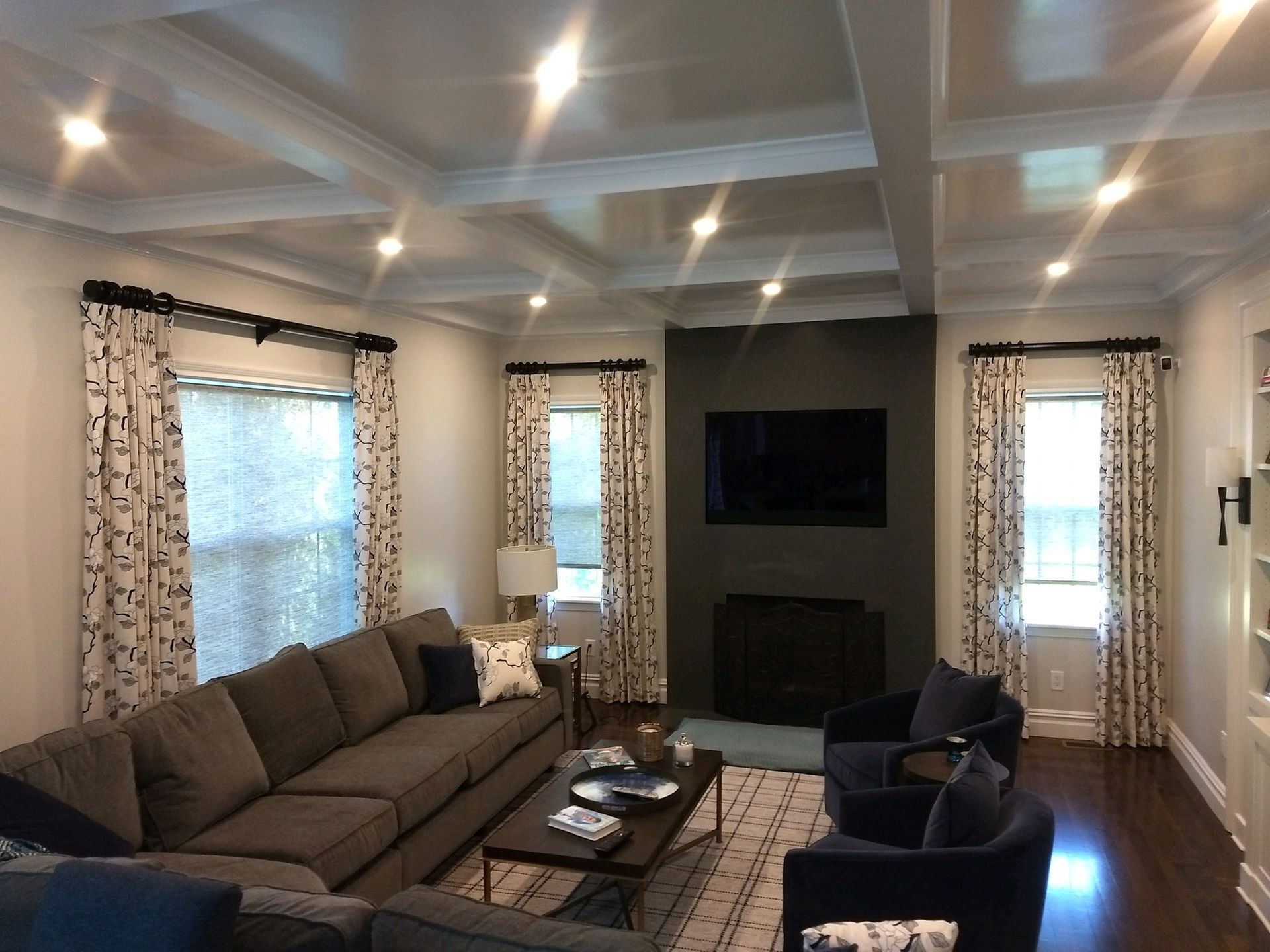 Living room with a gray sofa, dark accent chairs, and patterned curtains. Fireplace with TV on a dark wall.