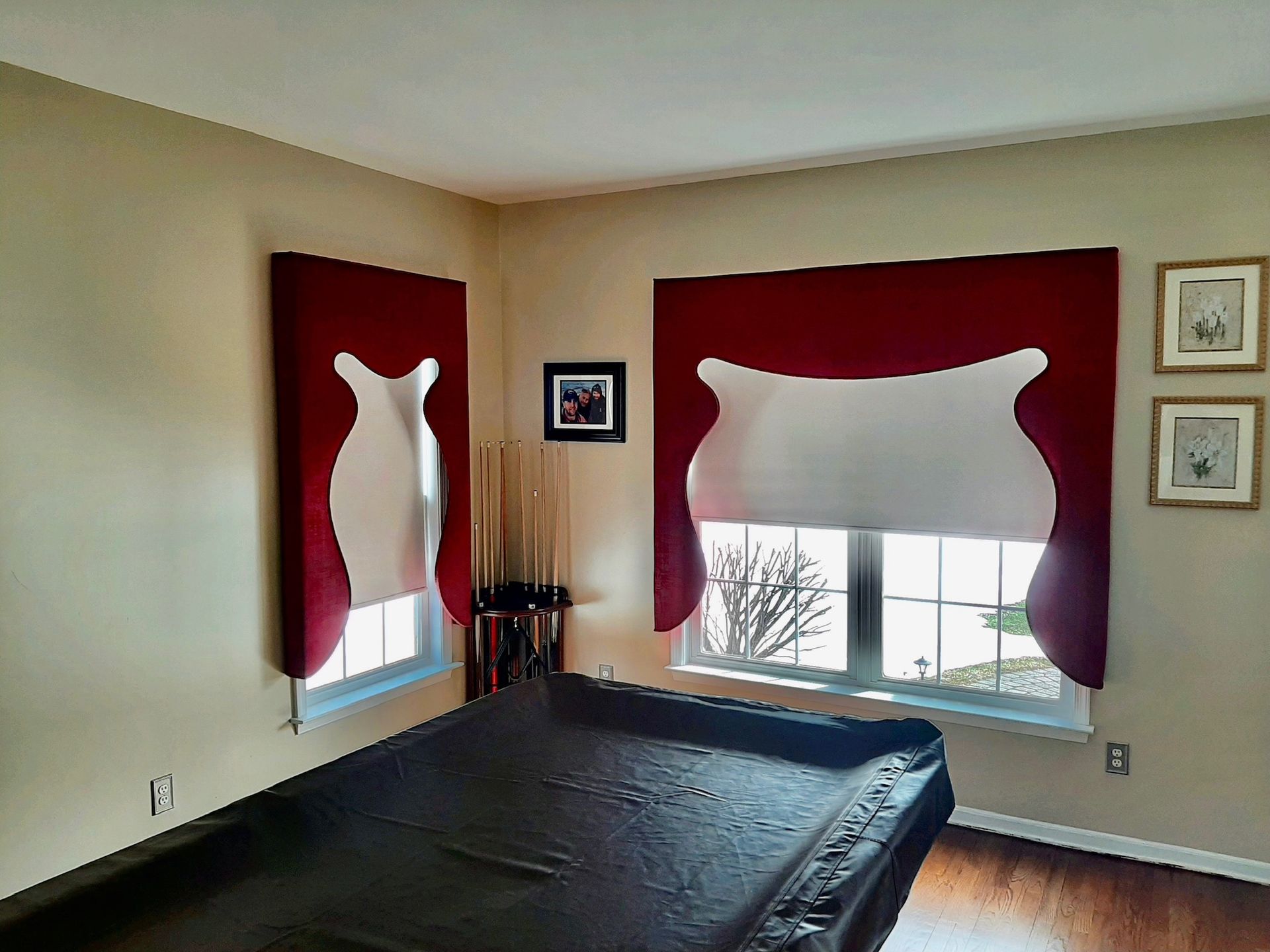 A room with a pool table, windows with red and white curtains, and light brown walls.