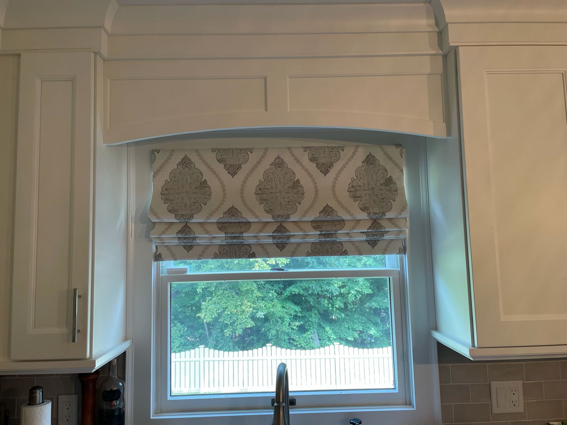 White kitchen cabinets frame a window with a patterned Roman shade, overlooking green foliage.