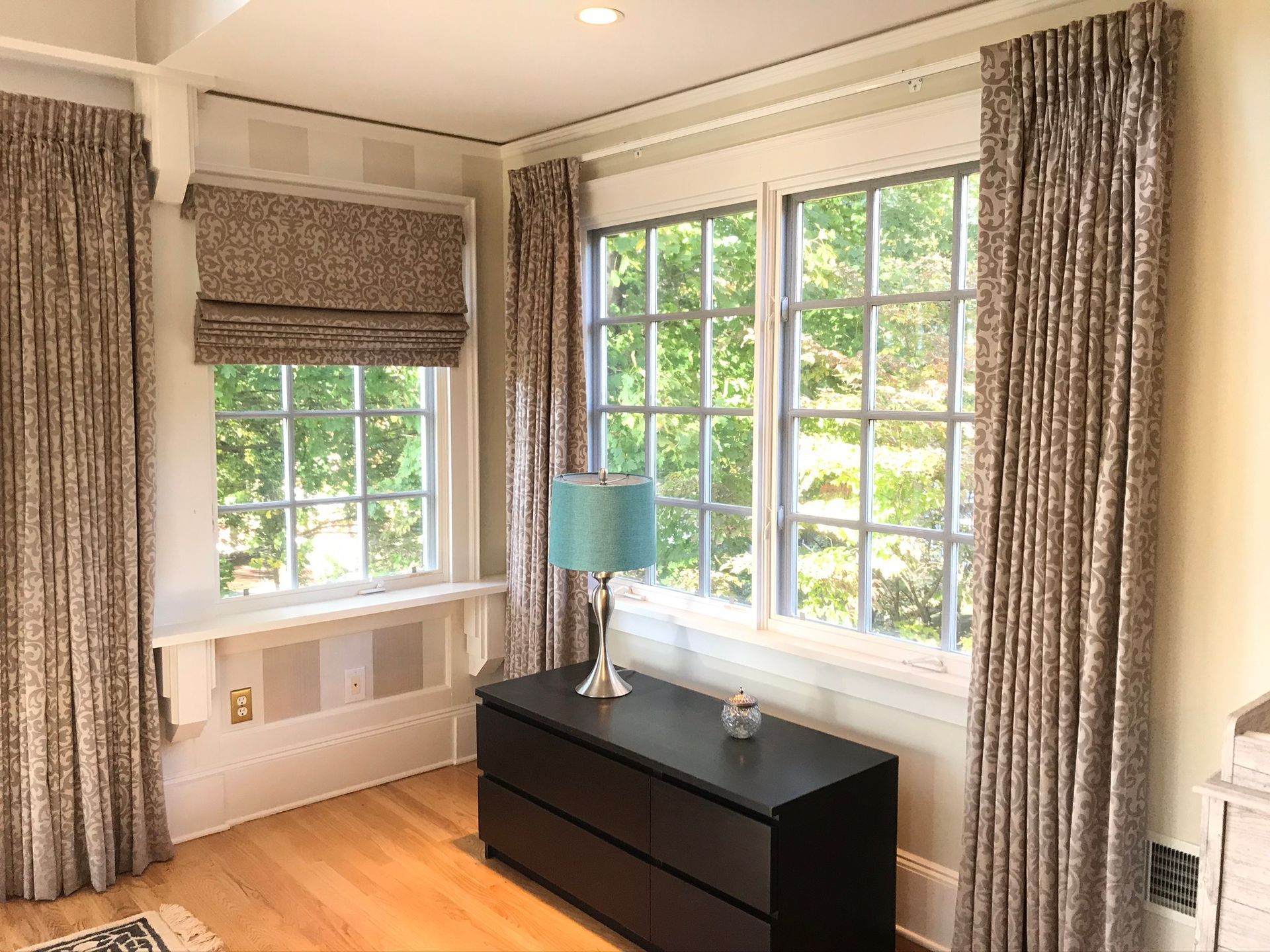 Room with windows, floral patterned curtains, and a dark dresser with a lamp.