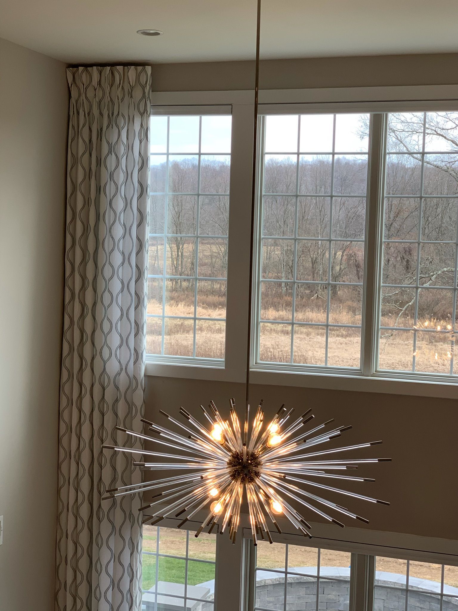 Modern chandelier hangs in front of a large window with a landscape view and patterned curtain.