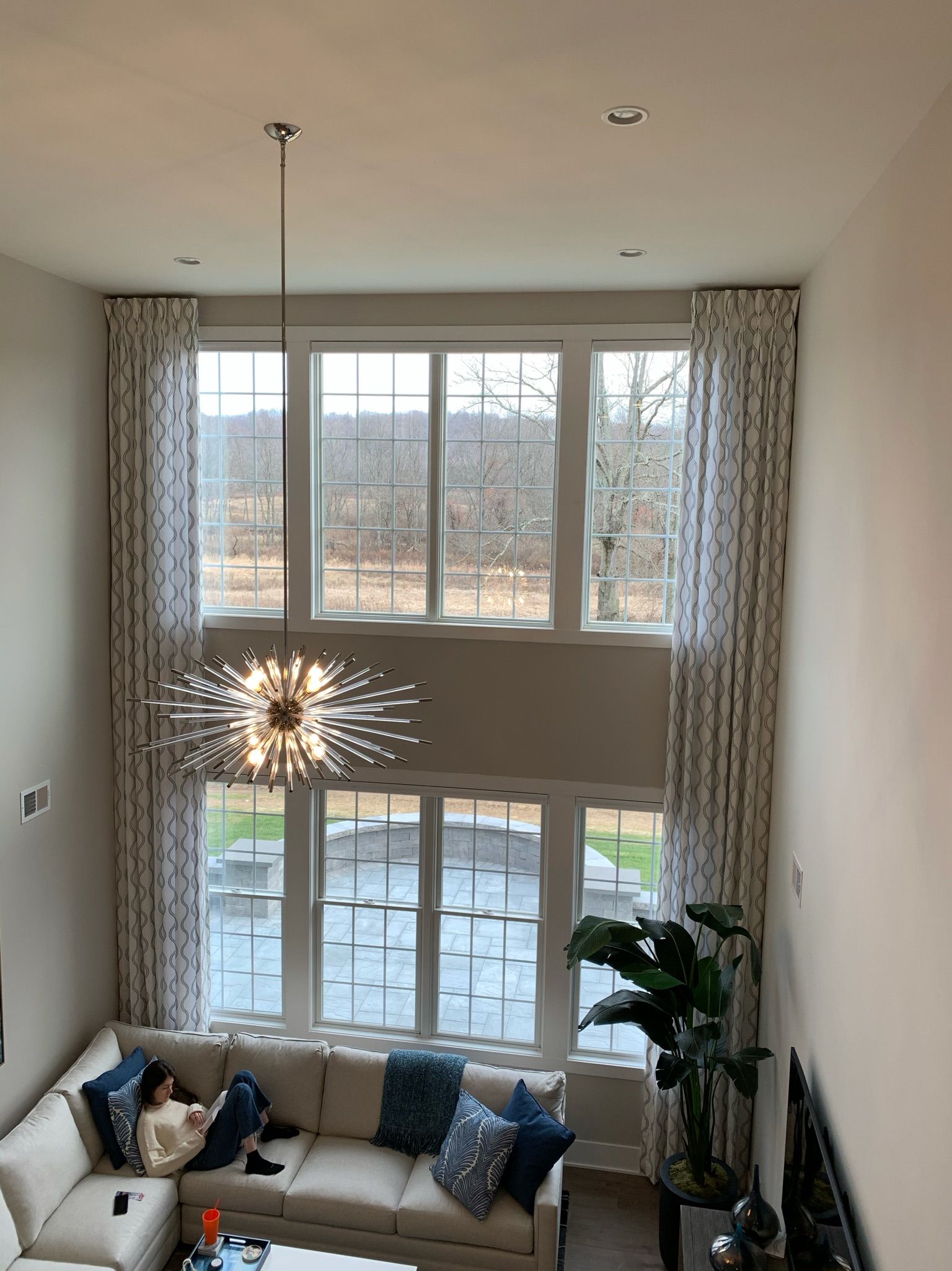 Woman relaxes on a sectional sofa below large, high windows with patterned curtains. A chandelier hangs in the center.