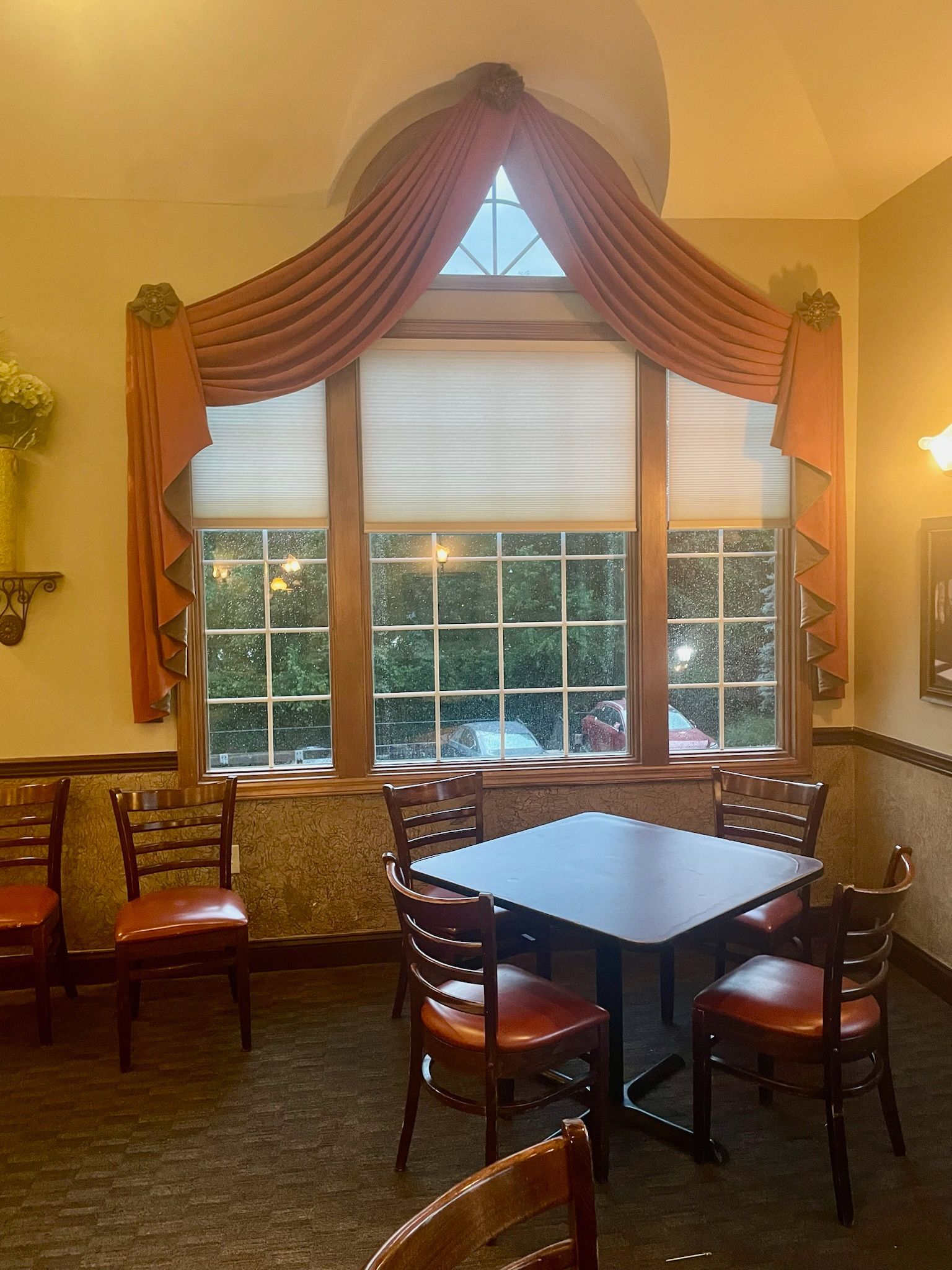 A restaurant interior with a square table, chairs, and a window with orange drapes.