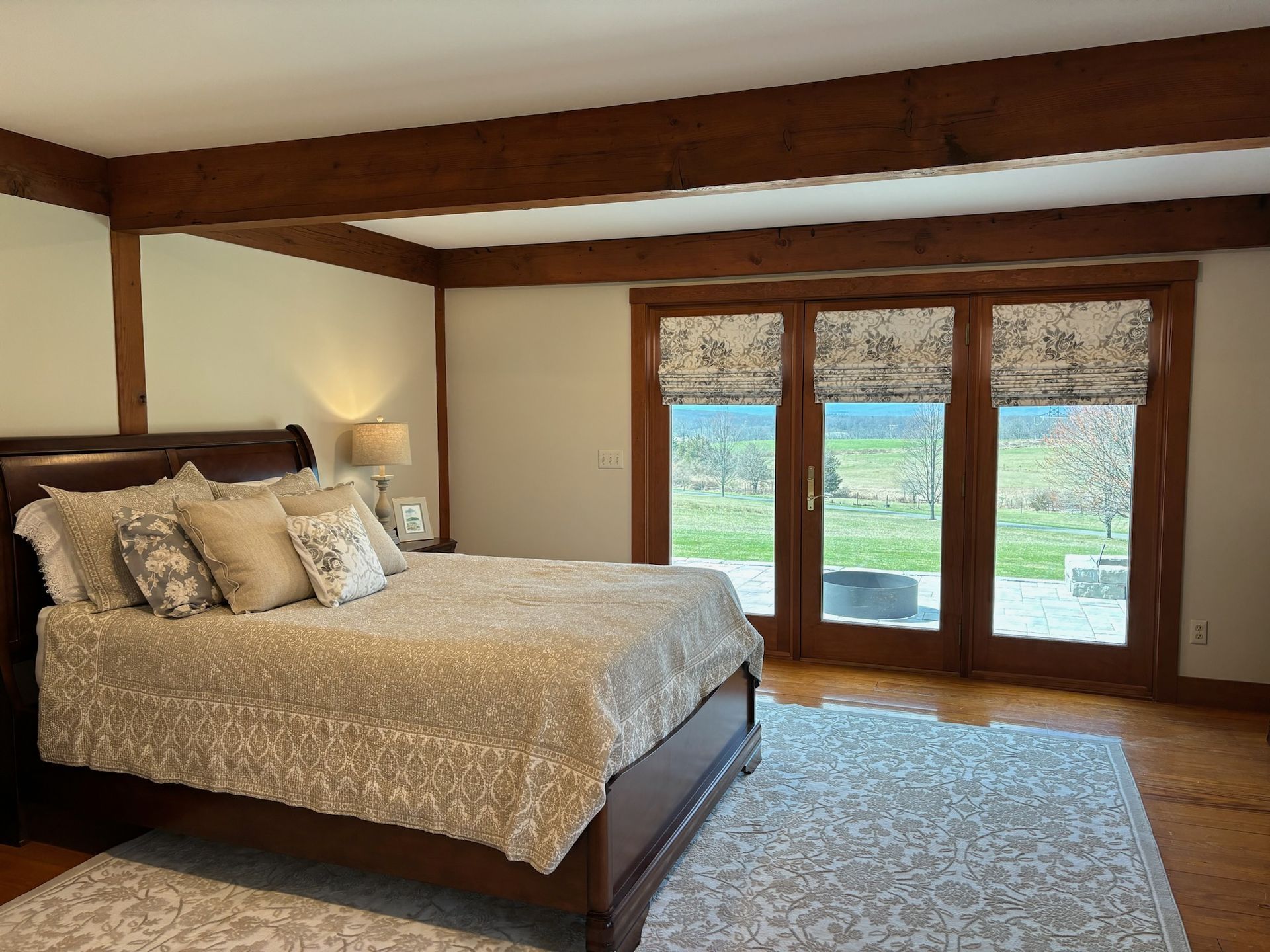 Bedroom with dark wood bed and beams, floral bedding, and French doors with shades overlooking a green lawn.
