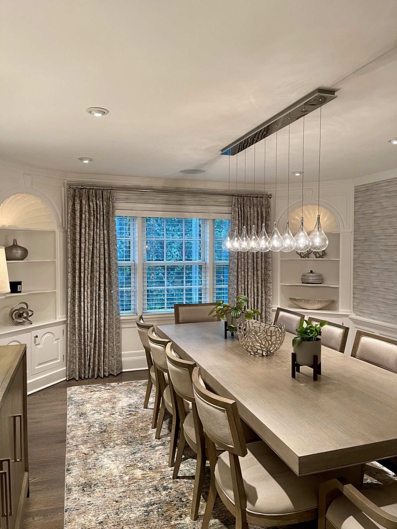Formal dining room with long wooden table and chairs, decorative curtains, built-in shelves, and chandelier.