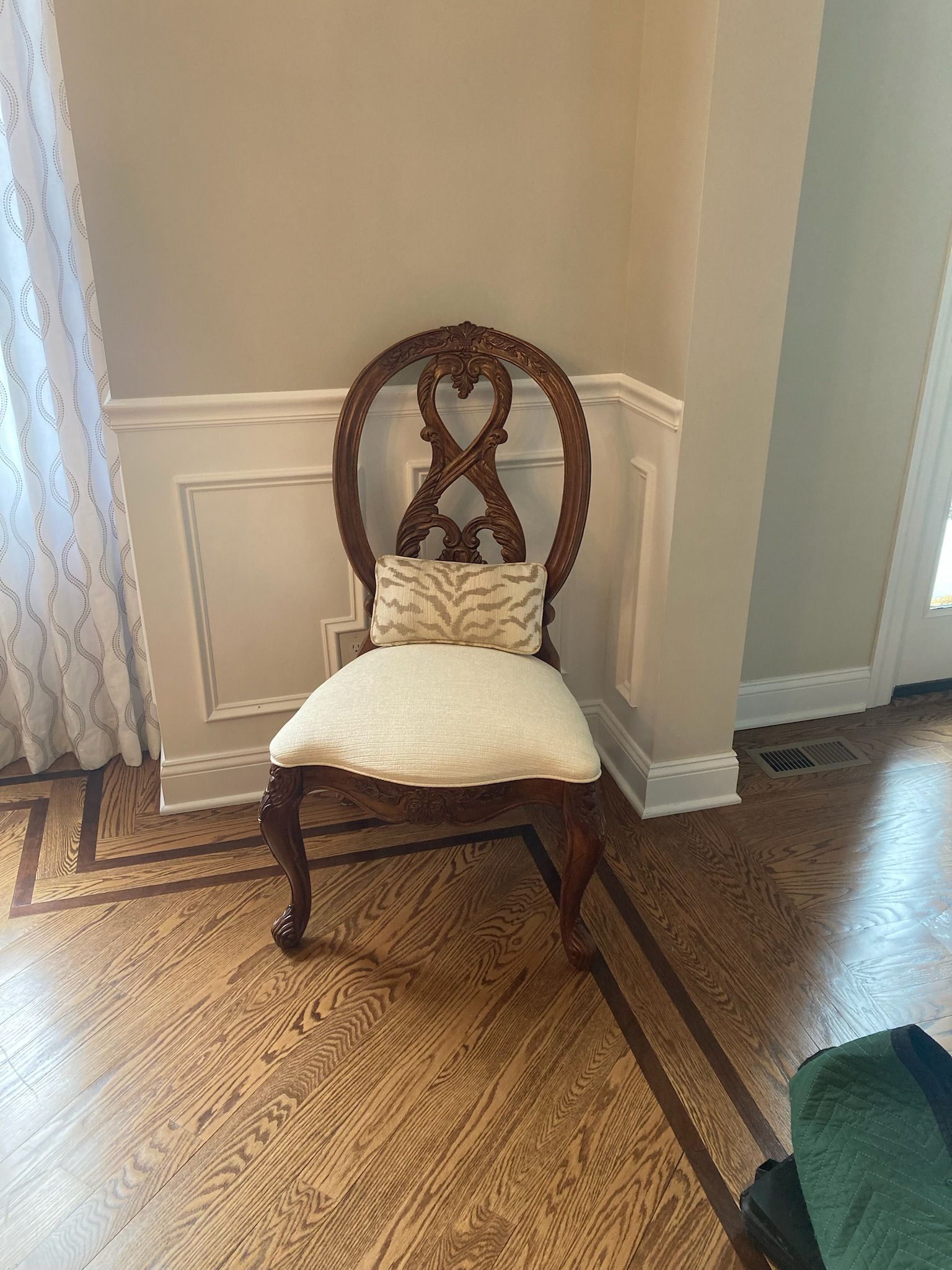 A decorative wooden chair with a cushion sits against a wall with molding. The floor is hardwood.