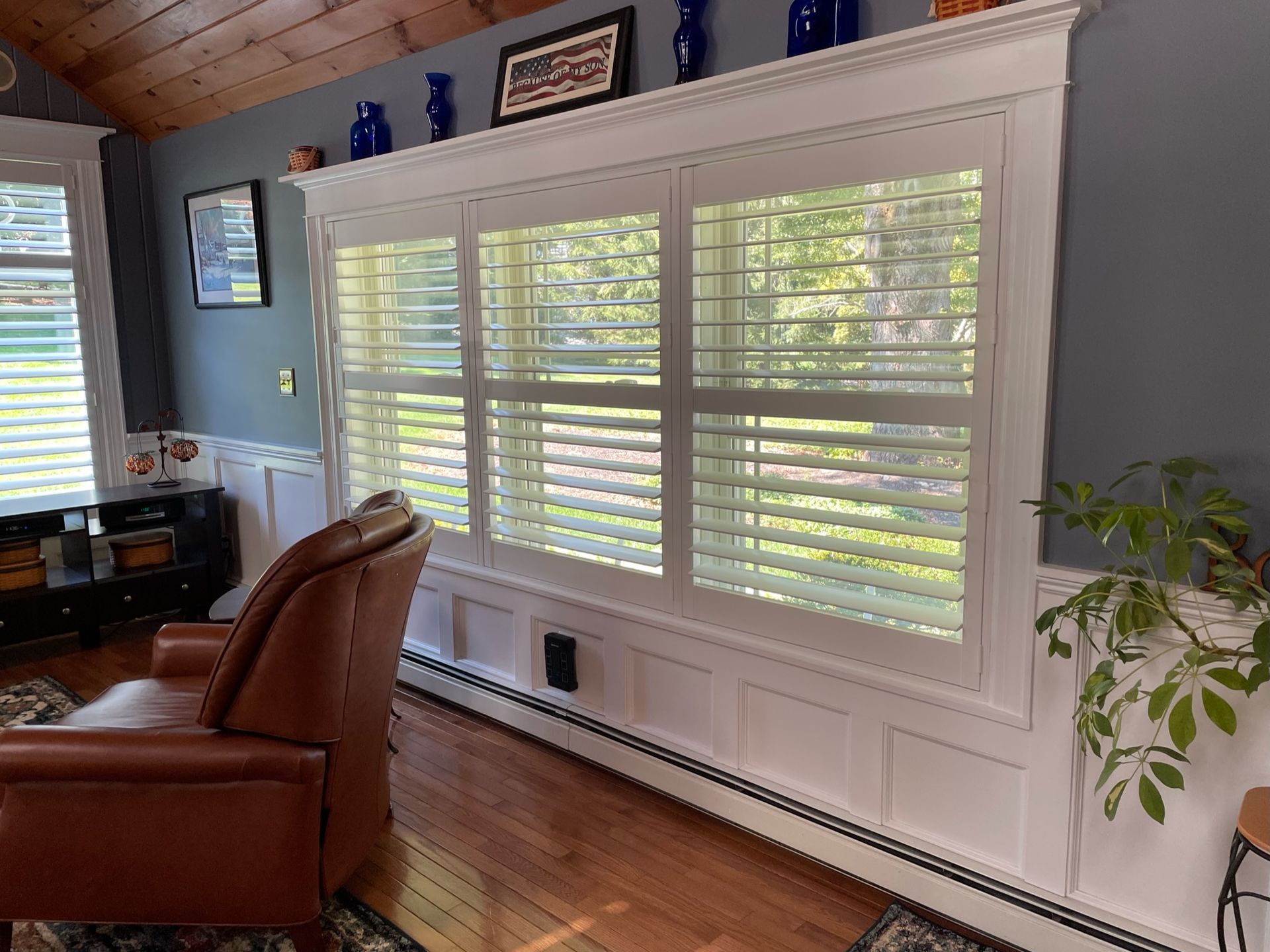 Interior with three-panel window with white shutters, brown leather chair, and wood flooring.