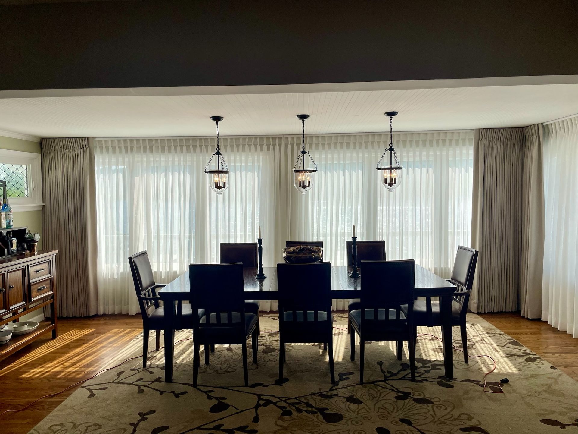 Dining room with dark table and chairs, three hanging lights, sheer curtains, and rug.