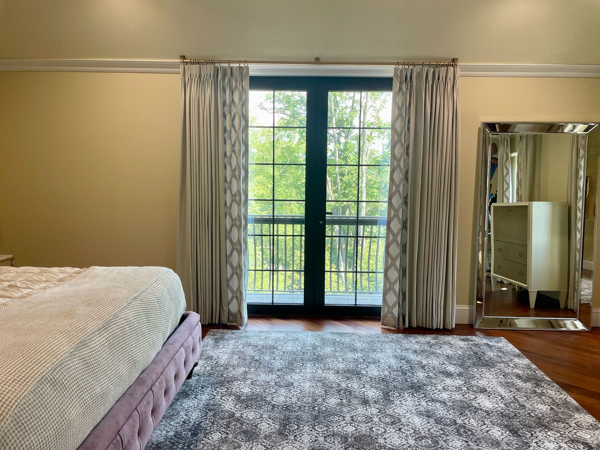 Bedroom with French doors, patterned curtains, large rug, and mirror. Light walls and hardwood floors.