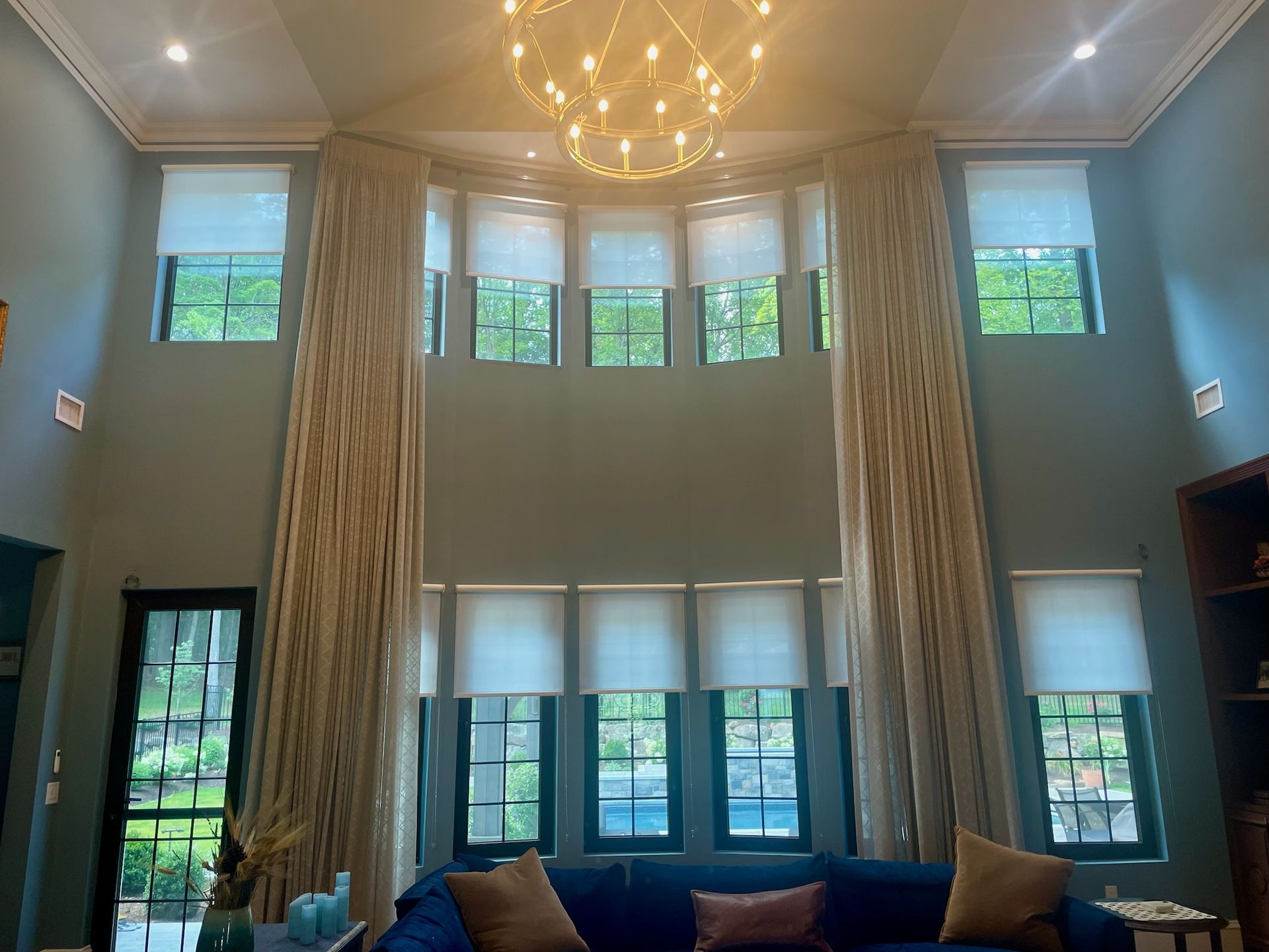 High-ceiling living room with large windows, light-colored curtains, and a blue sofa. A chandelier hangs above.