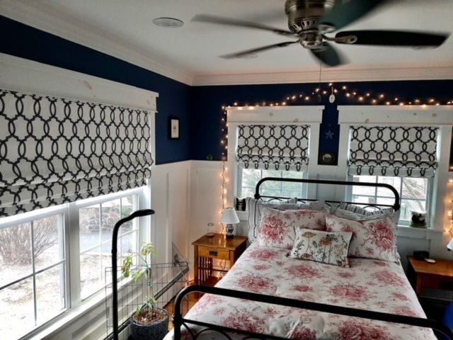 Bedroom with navy walls, white trim, patterned window shades, wrought iron bed, and string lights.