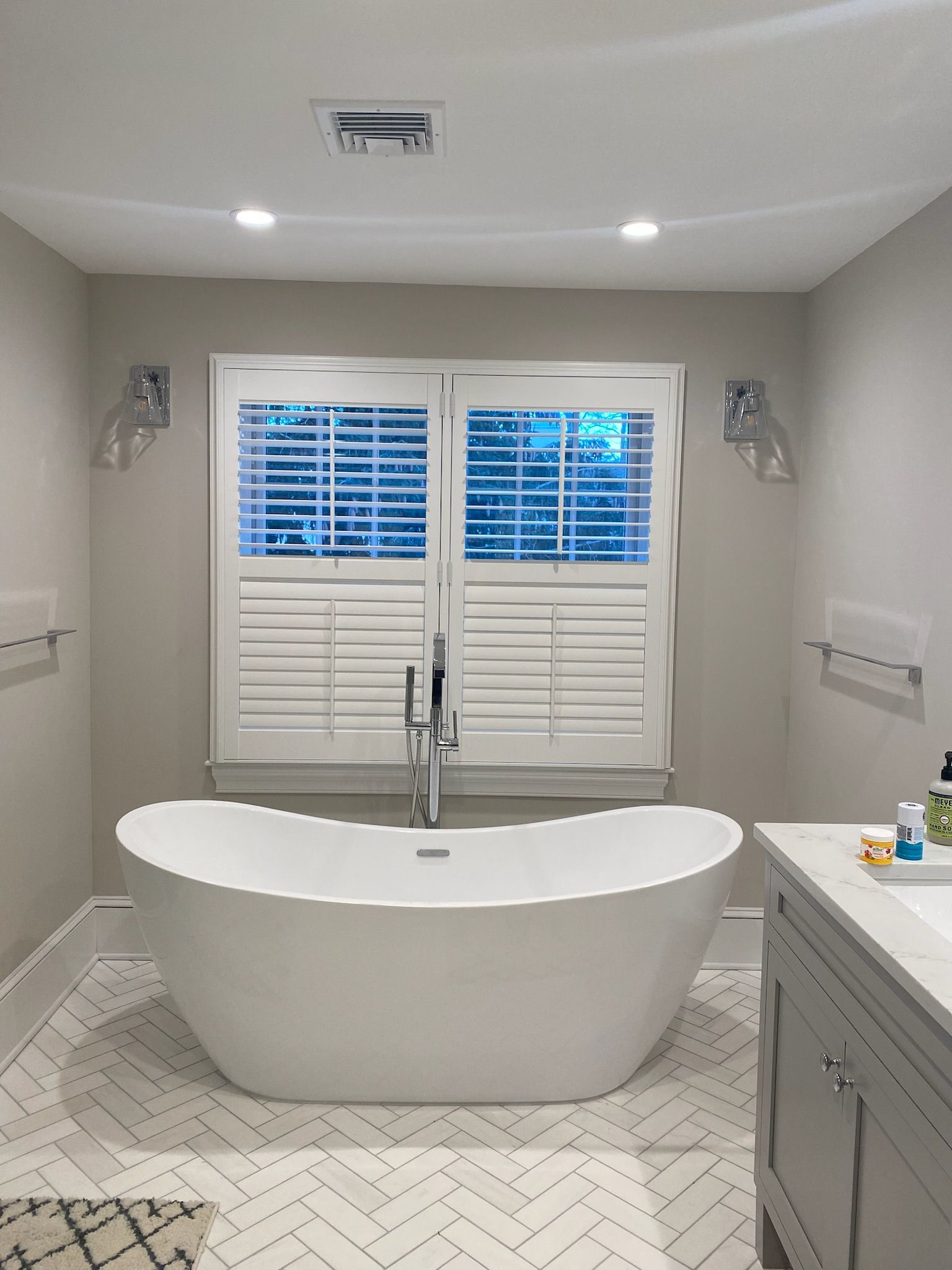 White bathtub in a modern bathroom with window shutters and herringbone tile flooring.