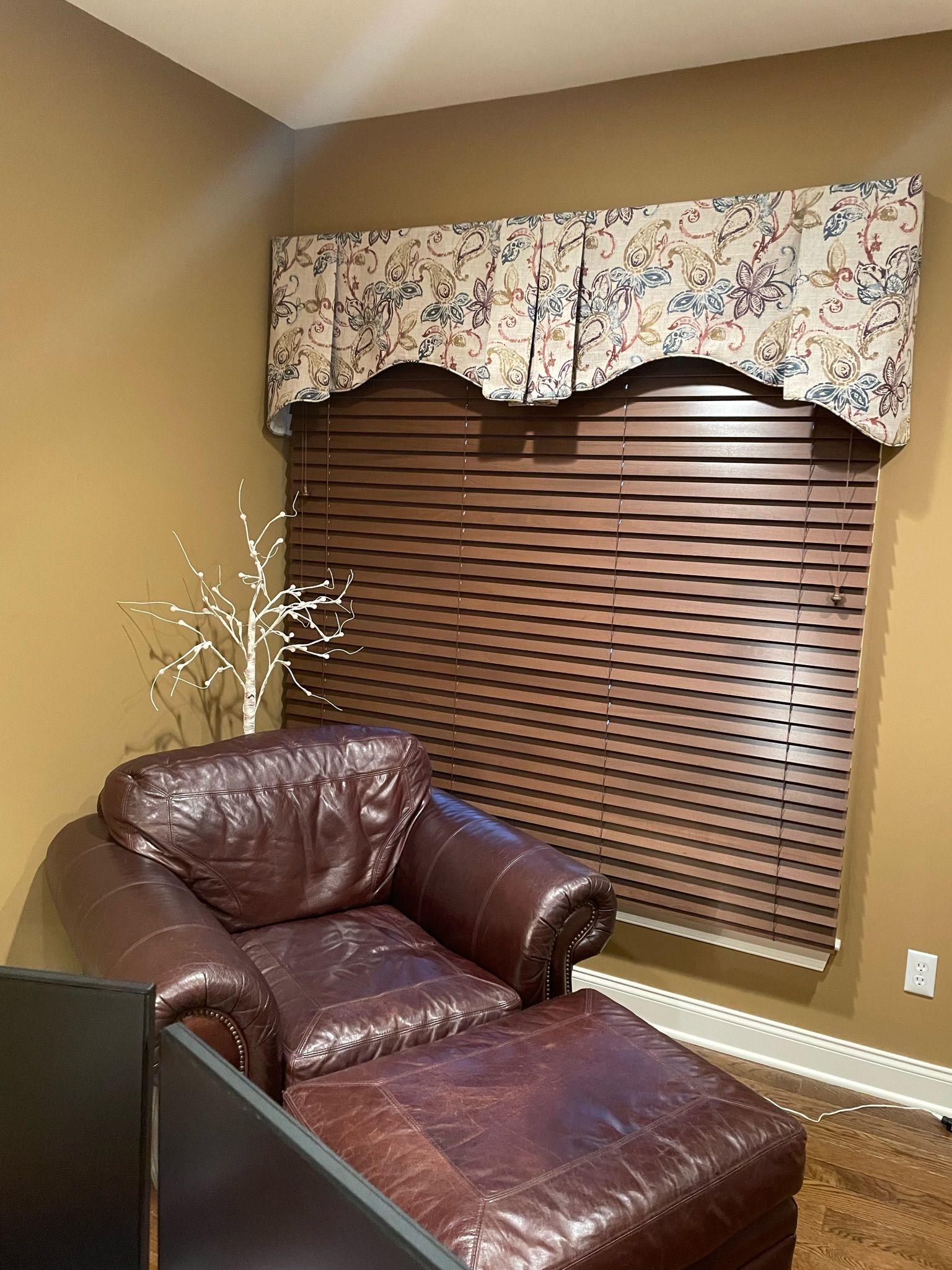Brown leather chair and ottoman by a window with blinds, a floral valance, and a decorative tree.