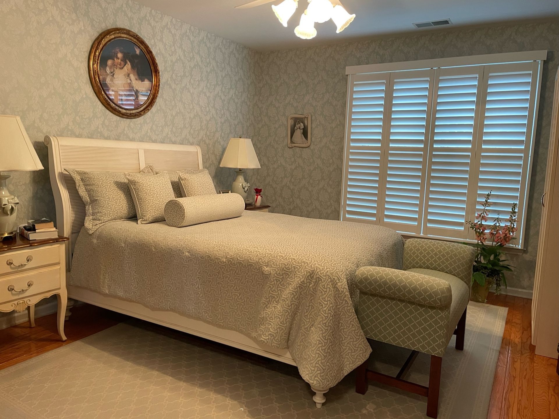 Cozy bedroom with a white bed, patterned wallpaper, and a small armchair near a window with white shutters.