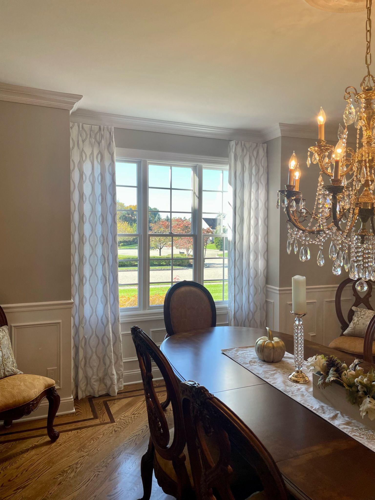 Formal dining room with bay window, table set for a meal, ornate chandelier, and light-colored patterned drapes.