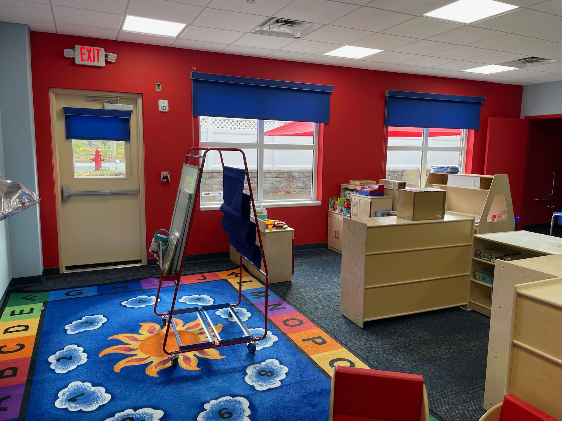 A colorful preschool classroom with a rug, easel, and wooden furniture; red walls, blue accents.