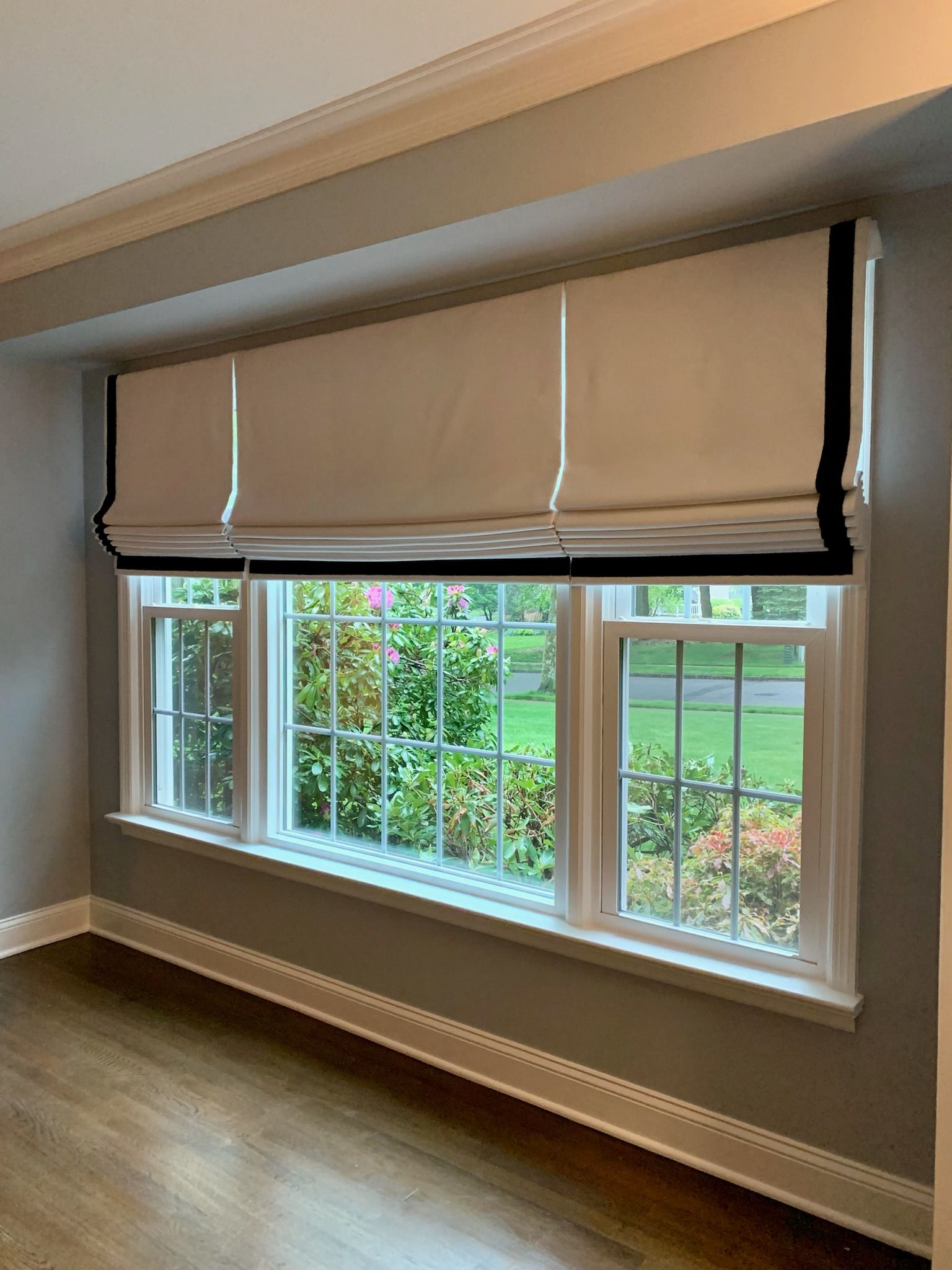 A window with a white and black roman shade, looking out onto a garden.