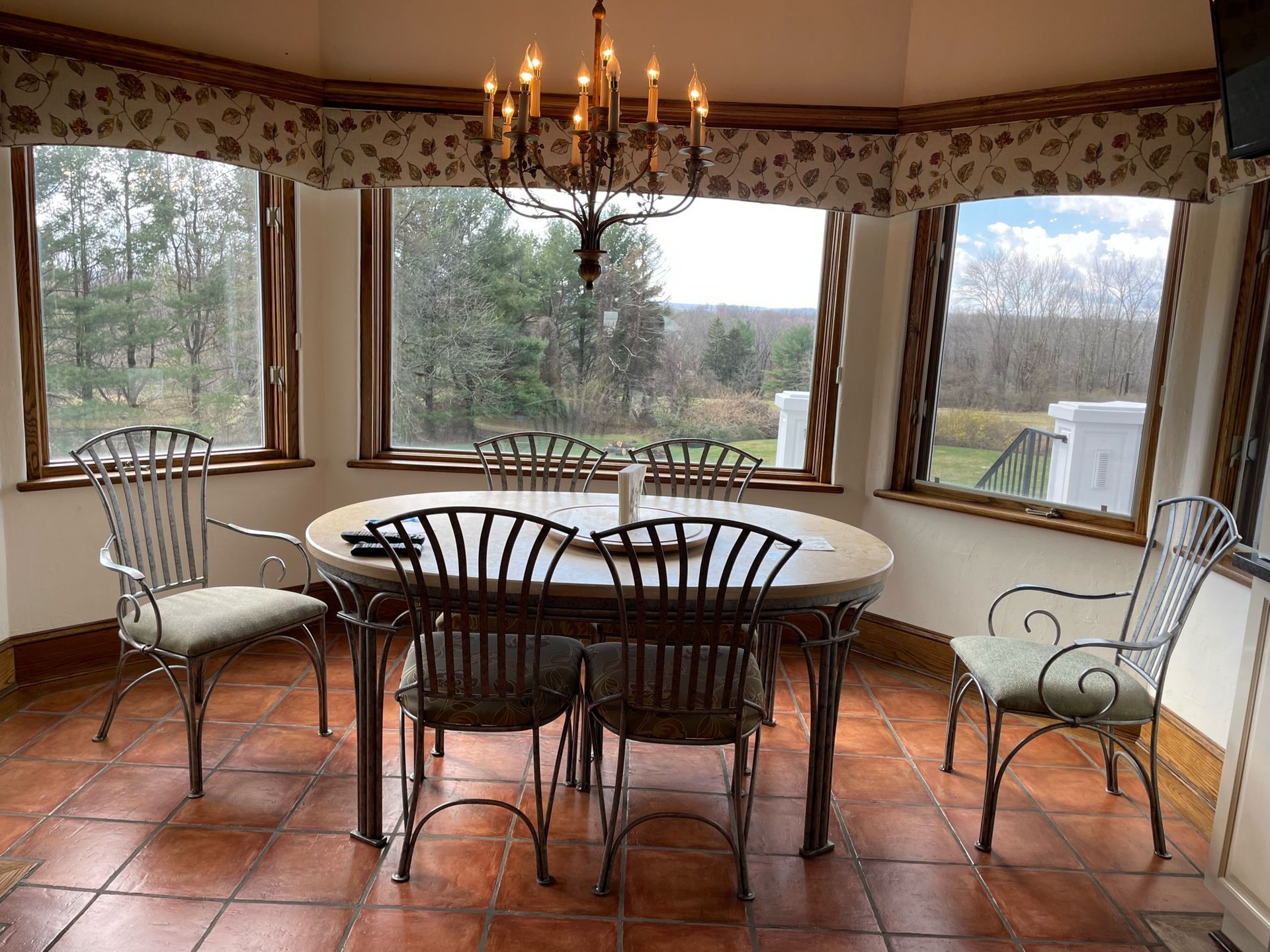Dining room with wrought iron furniture, oval table, and a chandelier; view of a wooded area.