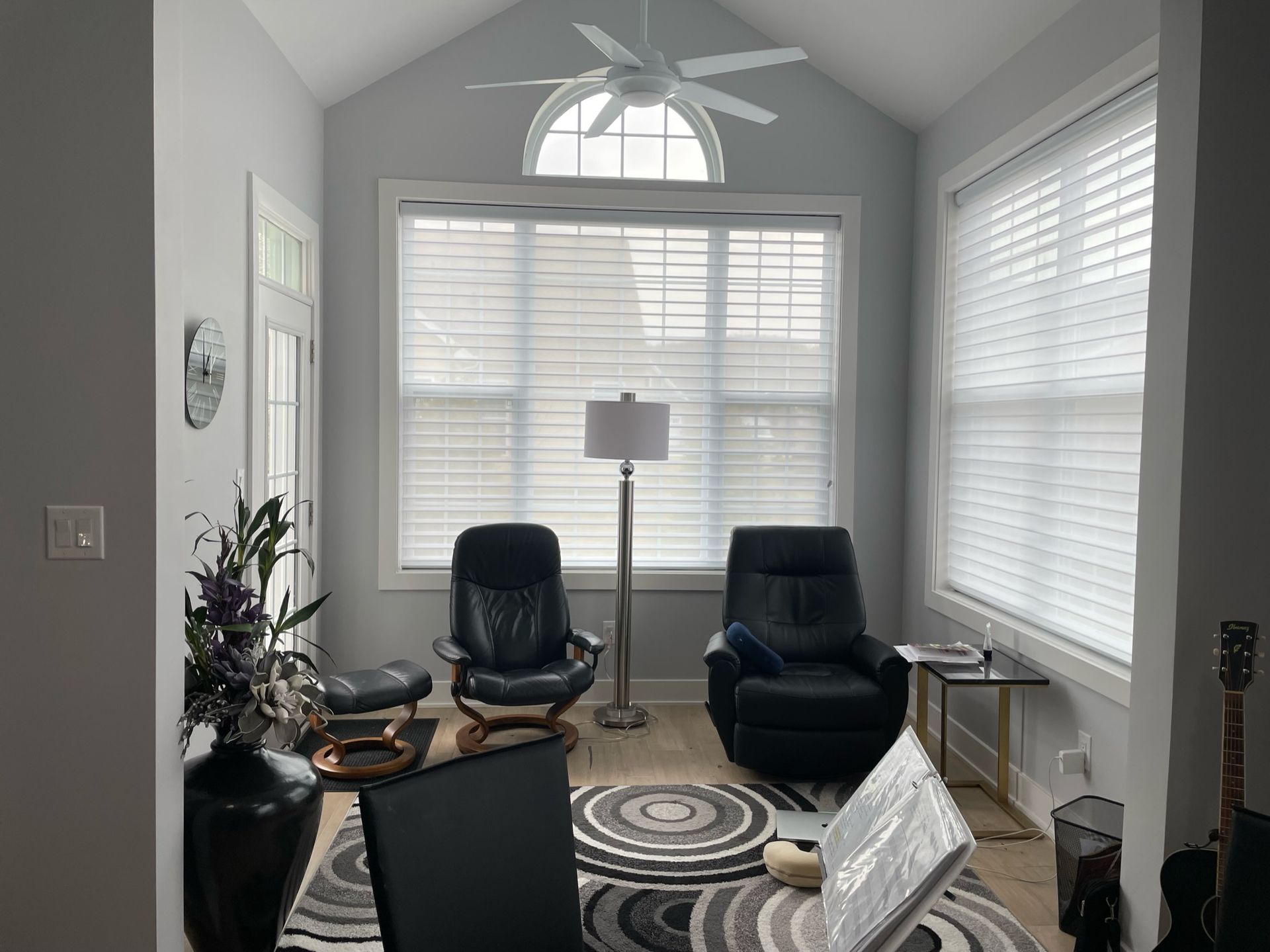 Cozy sunroom with black recliners, a floor lamp, and a patterned rug. Gray walls, arched window, and a ceiling fan.