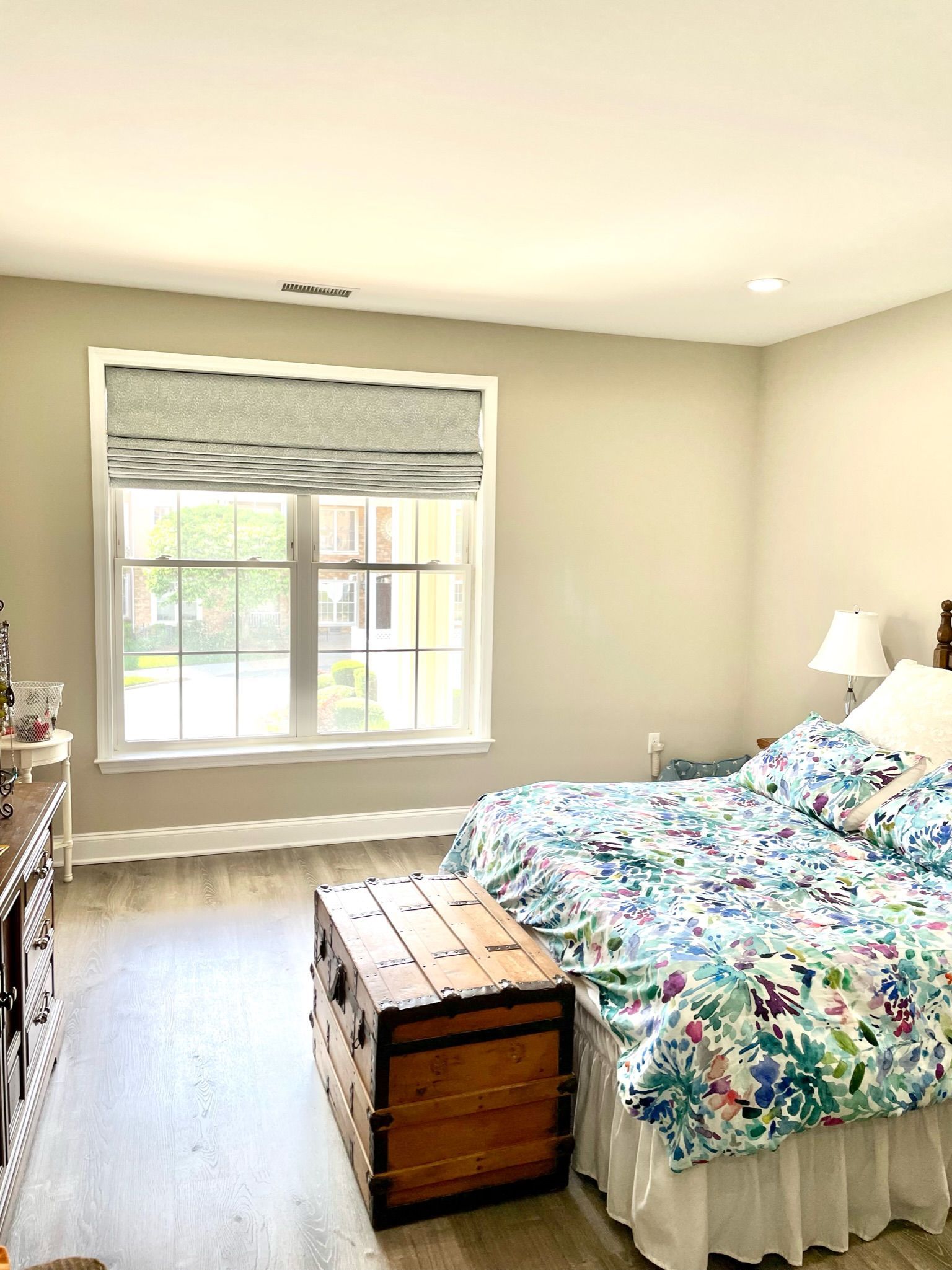 Bedroom with window, chest, bed with floral comforter, dresser, and light gray walls.