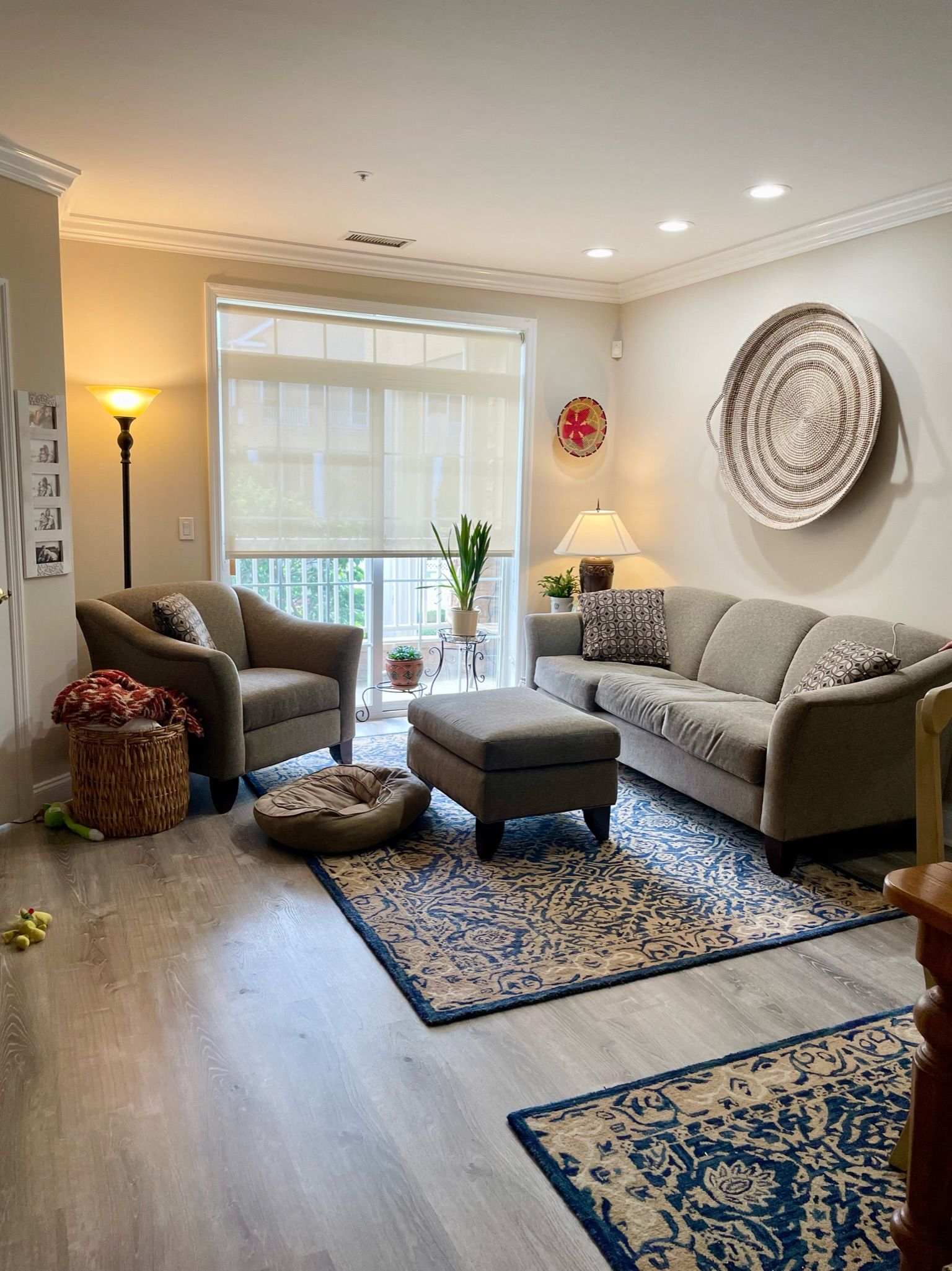 Living room with sofa, armchair, ottoman, rugs. Beige walls, grey flooring, light from window and lamps.