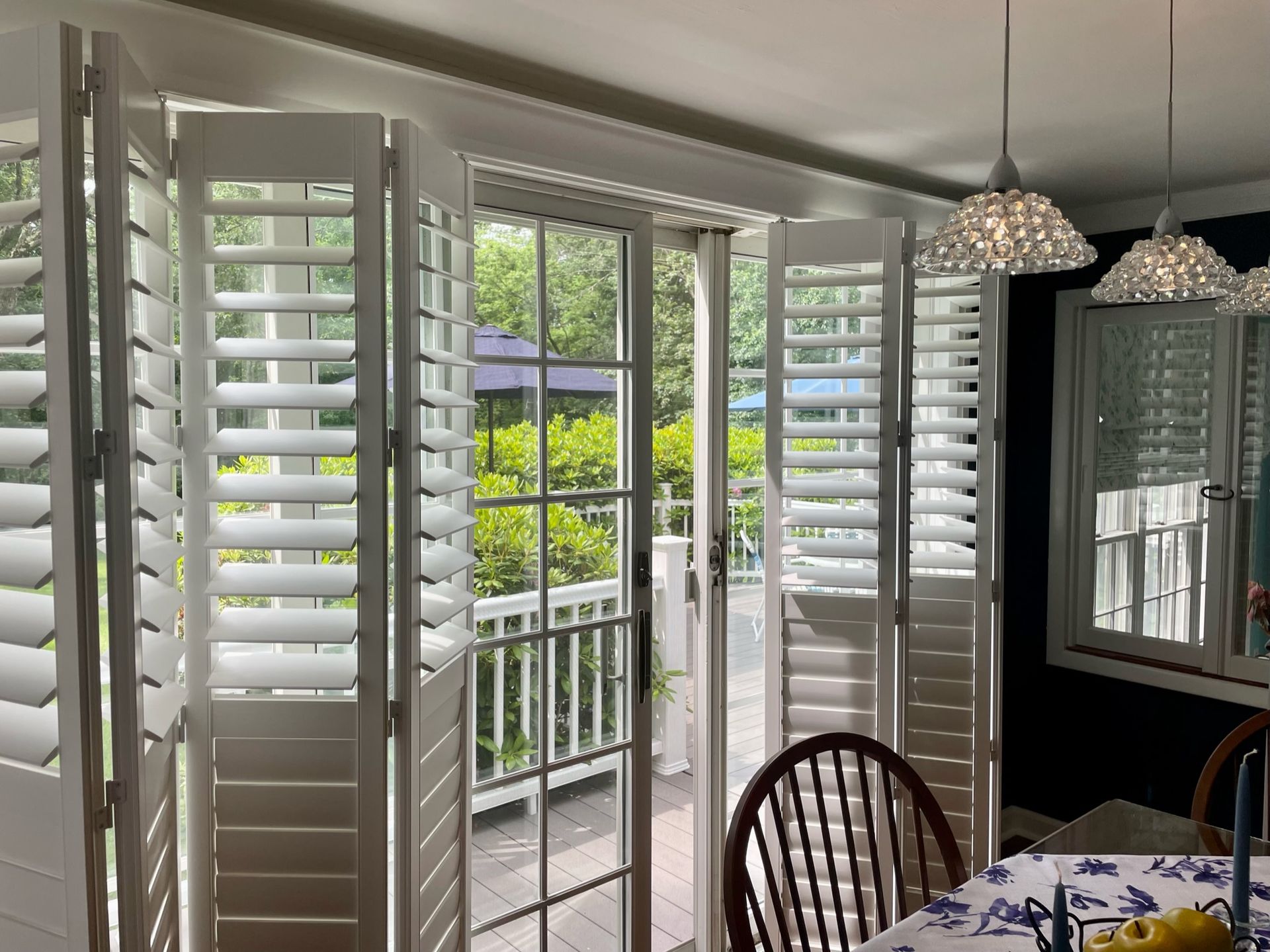 White shutters on a glass door and window with an outdoor view. Dining room table and chairs are visible.