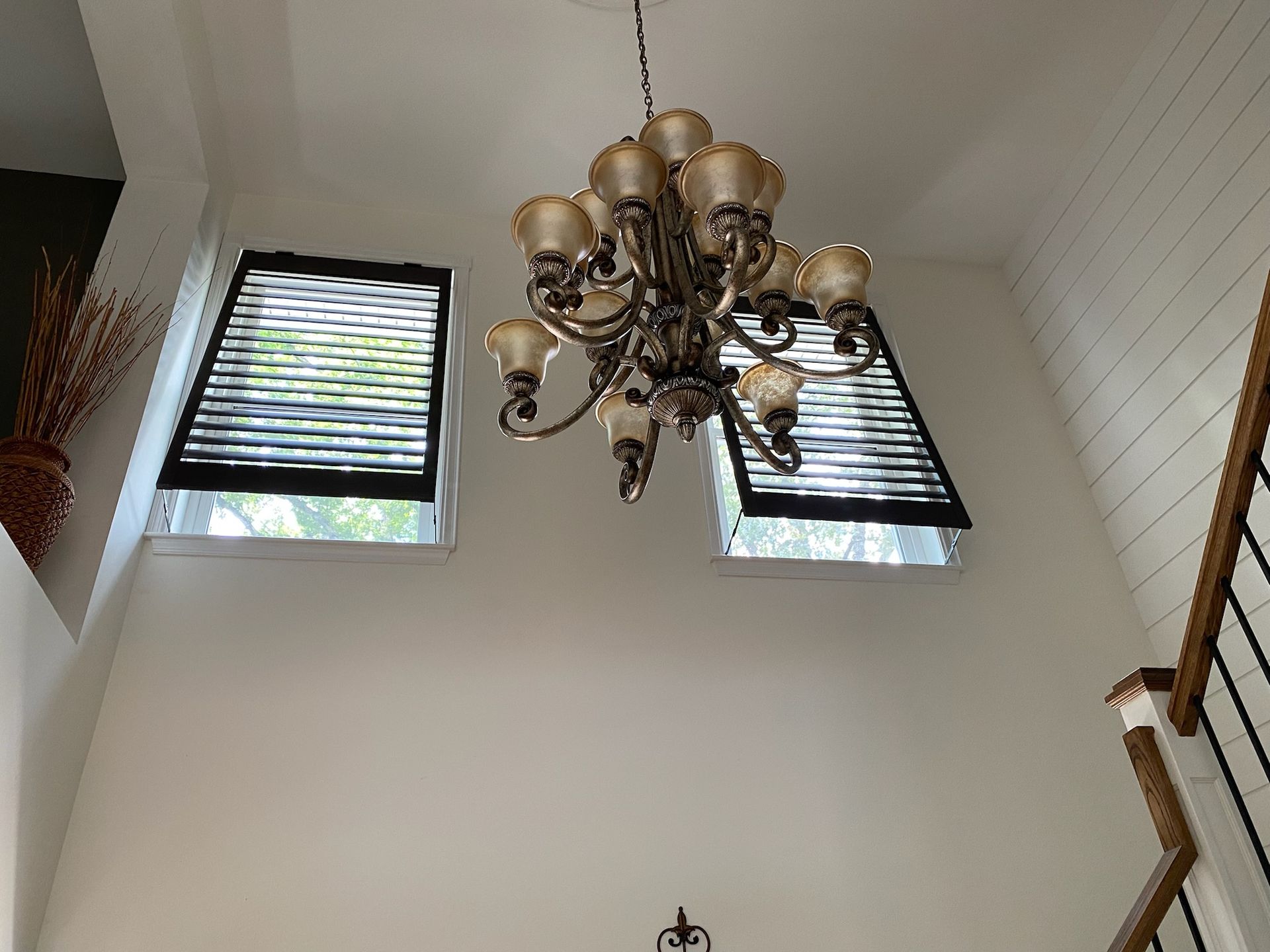 Chandelier hangs between two windows with open blinds. White walls, a staircase, and decorative flowers are visible.