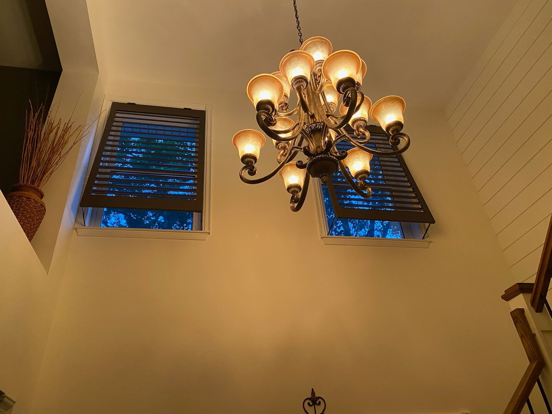 A chandelier hangs between two windows with shutters. Blue lights are visible through the windows.