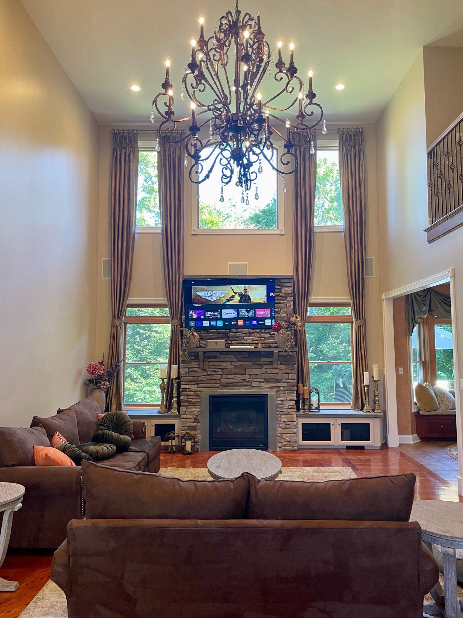 Living room with high ceilings, chandelier, stone fireplace, TV, and brown furniture.