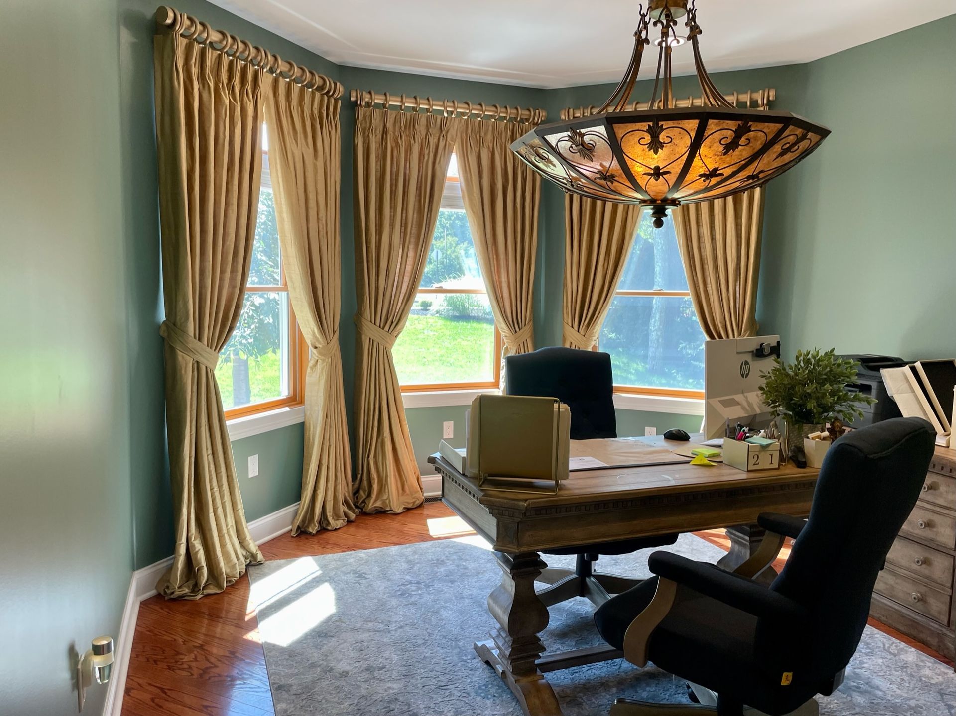 Home office with gold drapes, a rustic desk, leather chair, and ornate chandelier.