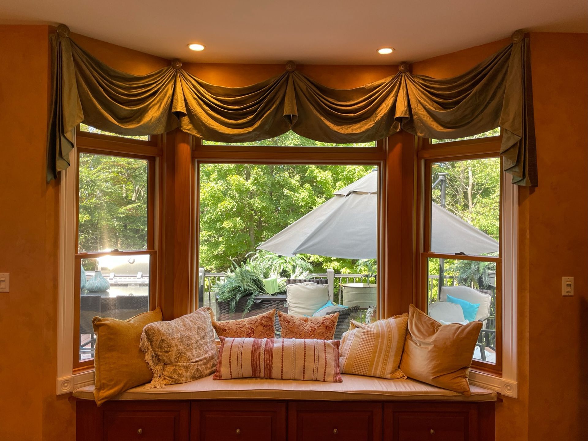 Bay window with valance, cushions on a bench, with a view of a patio and trees.