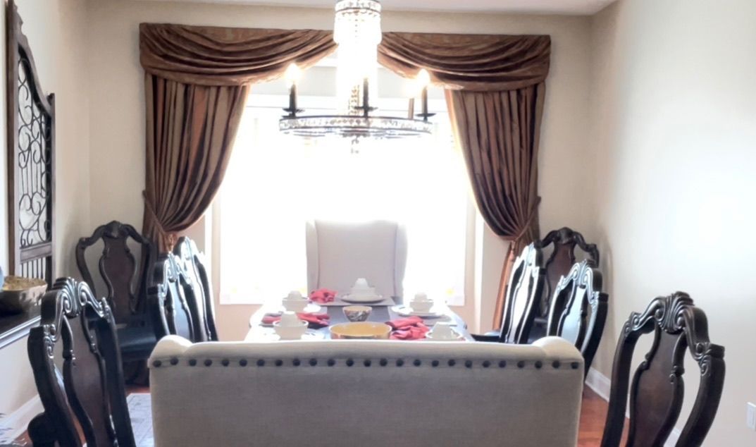 Dining room with ornate dark chairs, light-colored table, brown drapes, and chandelier.