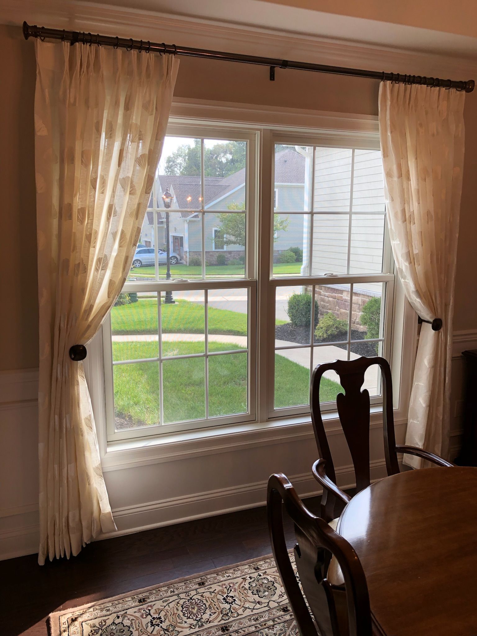 Window with sheer cream-colored curtains framing a view of a suburban street. Brown curtain rods and dining chair visible.