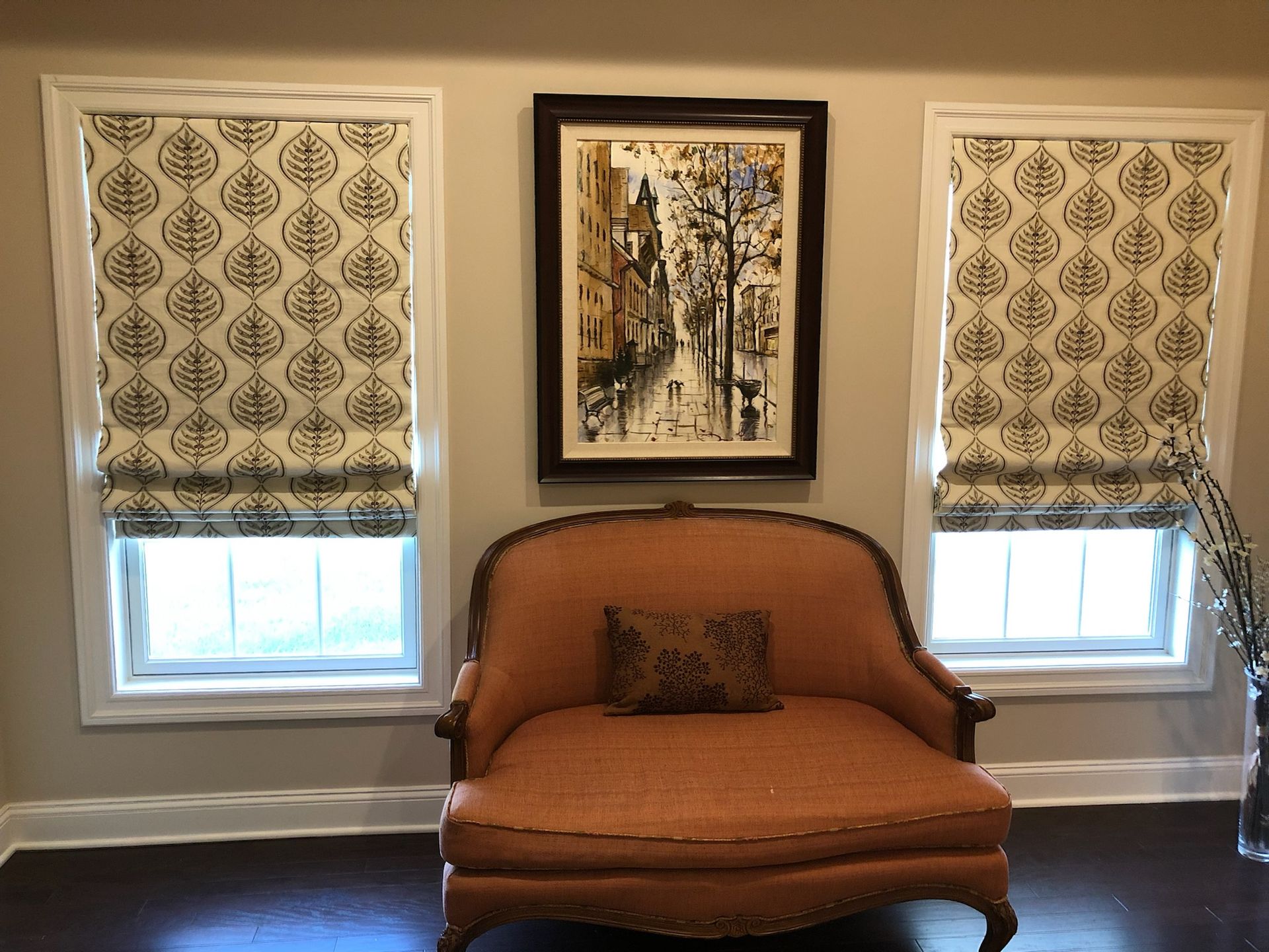 Two windows with patterned shades flank a chair beneath a framed painting, on a wood floor.