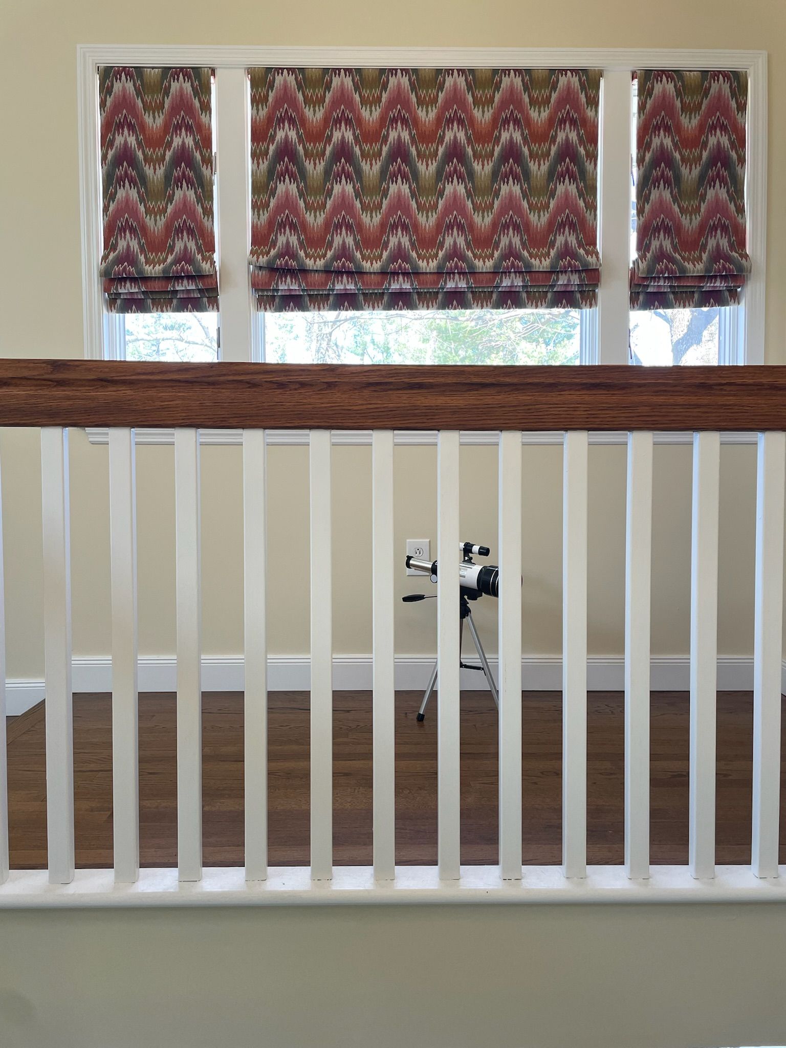 White railing, three windows with patterned Roman shades, neutral walls, wooden floor.