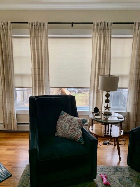 Living room with dark green armchair, beige curtains, roller shades, and small round table.