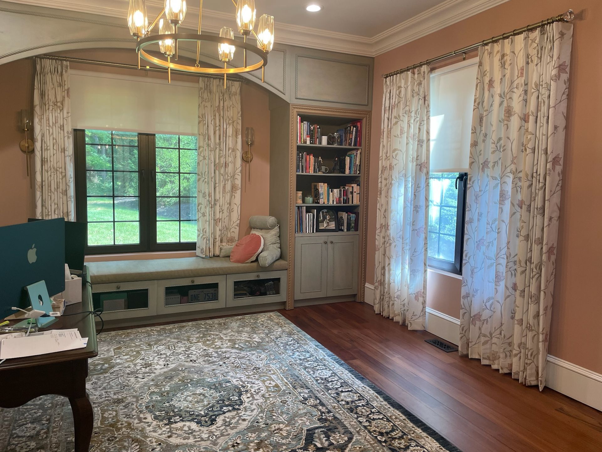 Room with window seat, bookshelves, and desk; soft pink walls, floral curtains, and an area rug.
