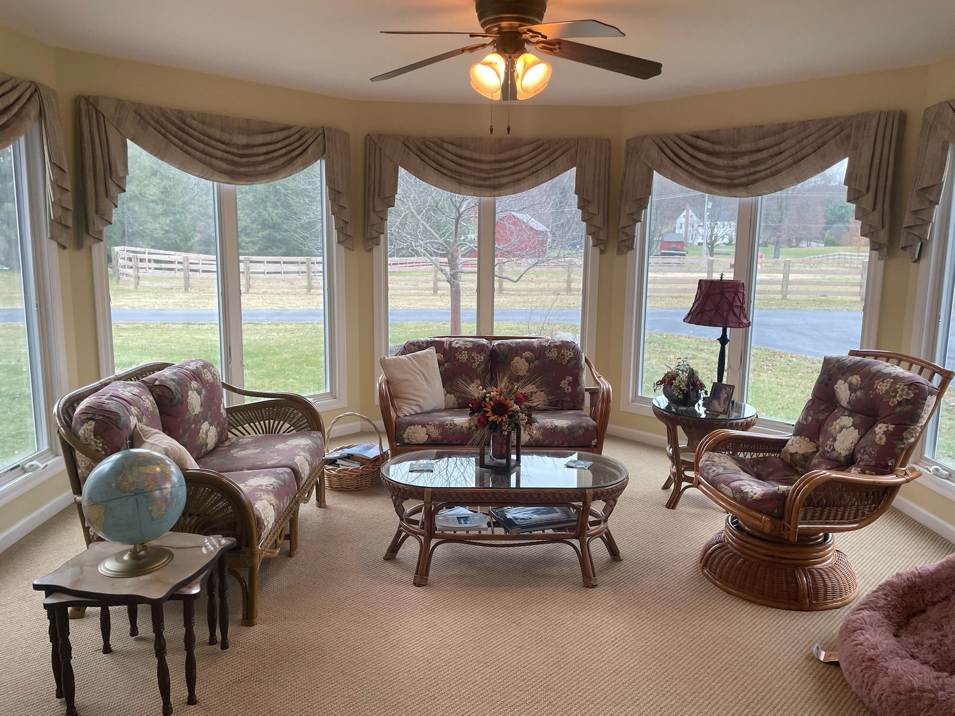 Sunroom with floral-patterned furniture, coffee table, and decorative drapes overlooking a green yard.