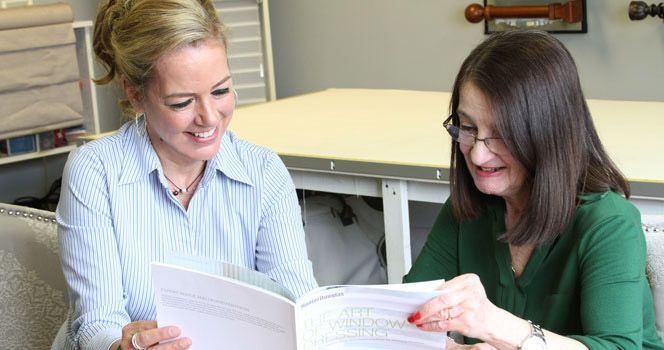 Two women, blonde and brunette, smiling while looking at a book in a room with samples.