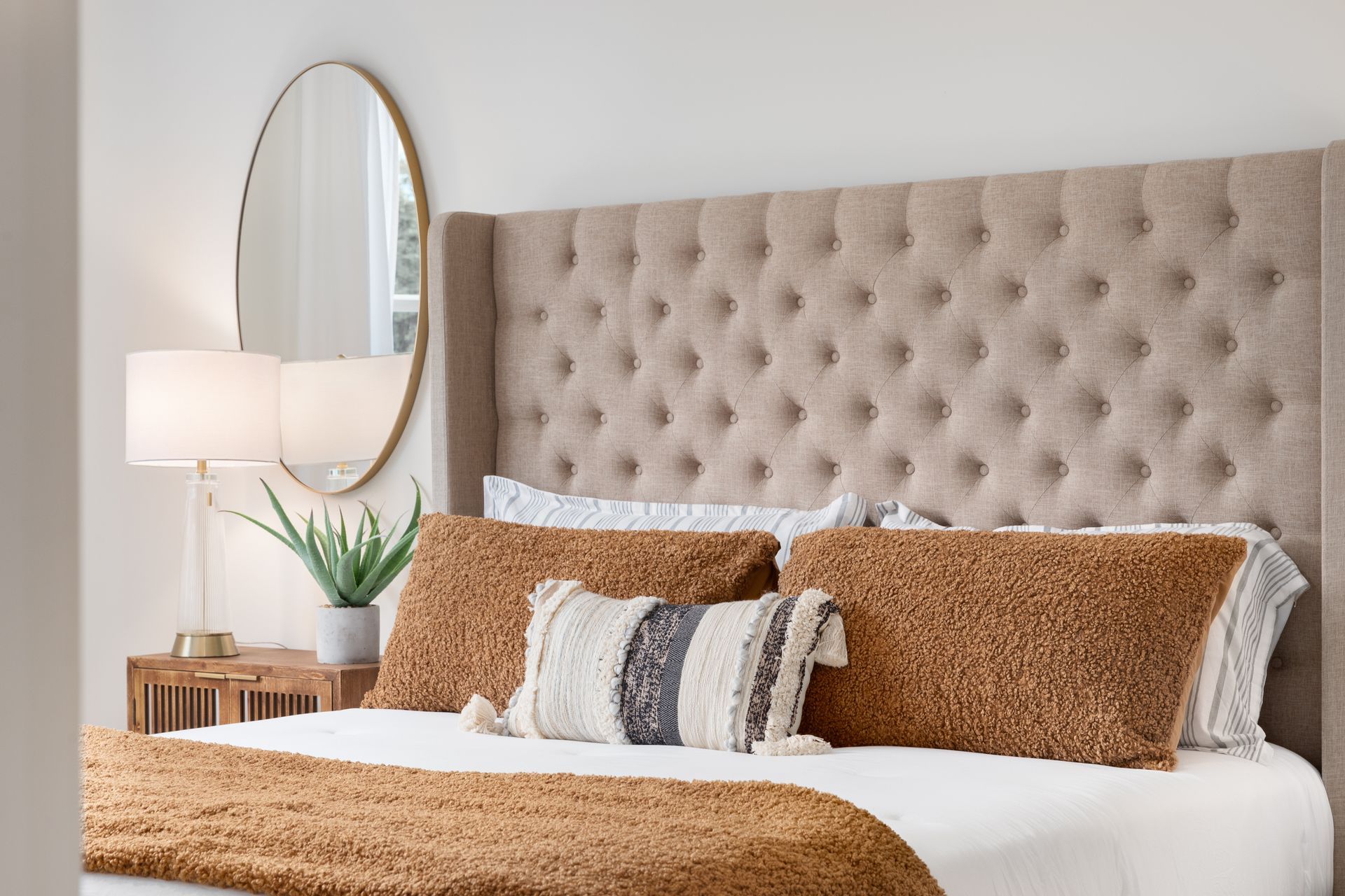 Bedroom with tufted headboard, bed, pillows, gold mirror, lamp, and wooden nightstand.