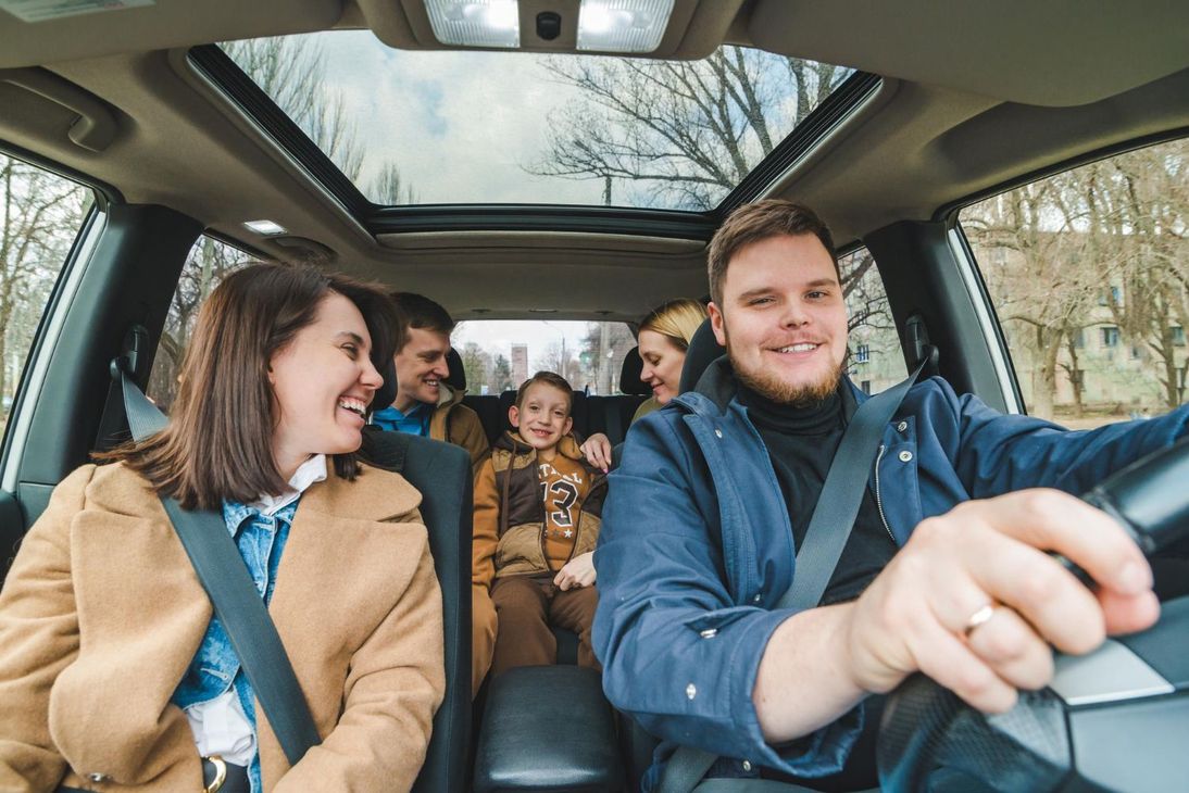 Family smiling in a car, with a driver in front and three passengers in the backseat.
