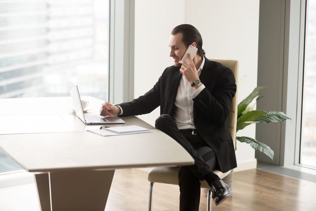 Man in a black suit works on a laptop while talking on a phone at a bright office desk.
