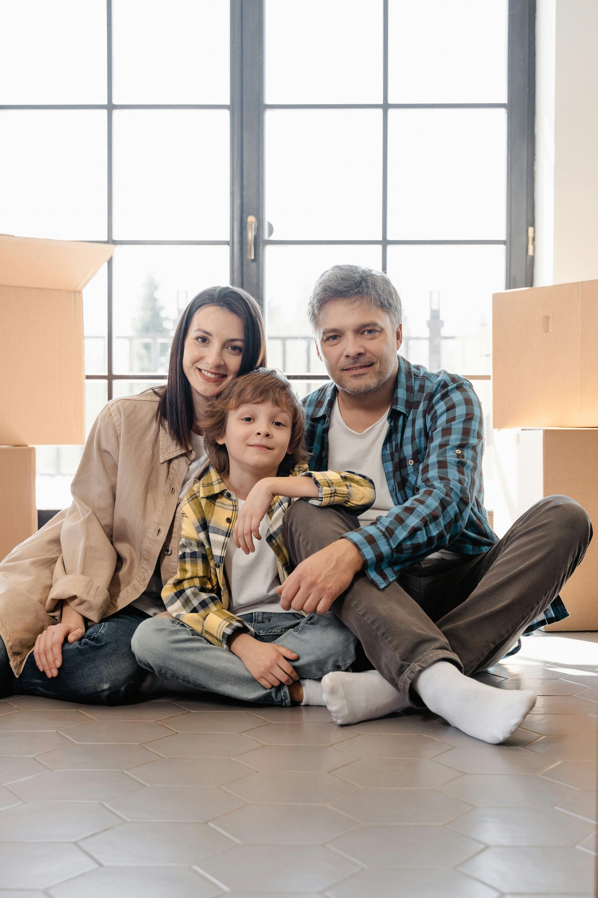 Family sitting on the floor by a large window in a bright living room with moving boxes