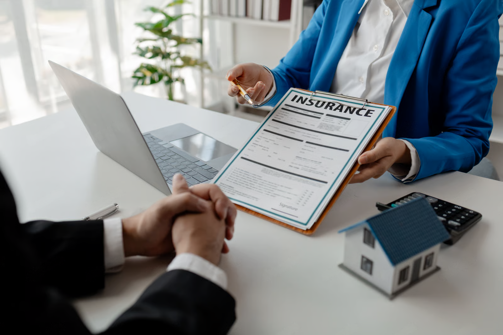 Insurance agent showing policy form across a desk in a bright office, with a laptop and small house model.