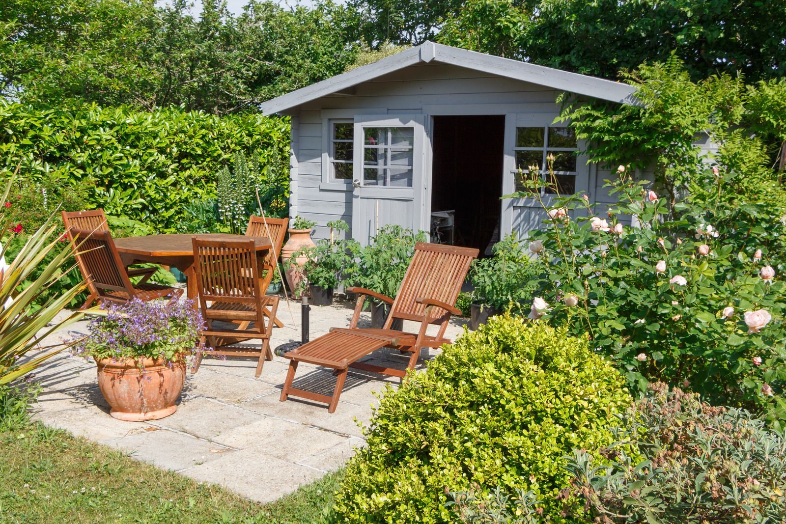 Garden patio with wooden chairs and table beside a small white shed, surrounded by lush greenery