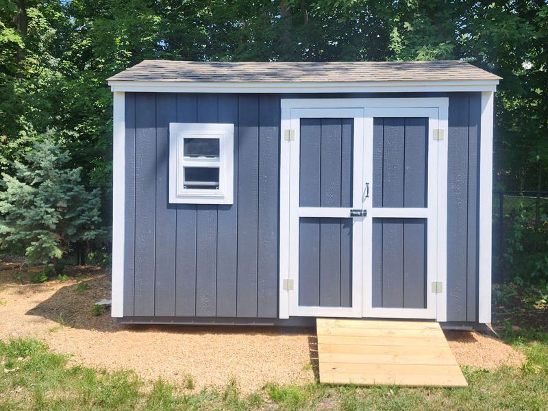 Small gray storage shed with white trim and double doors, set outdoors on a gravel path surrounded by trees.